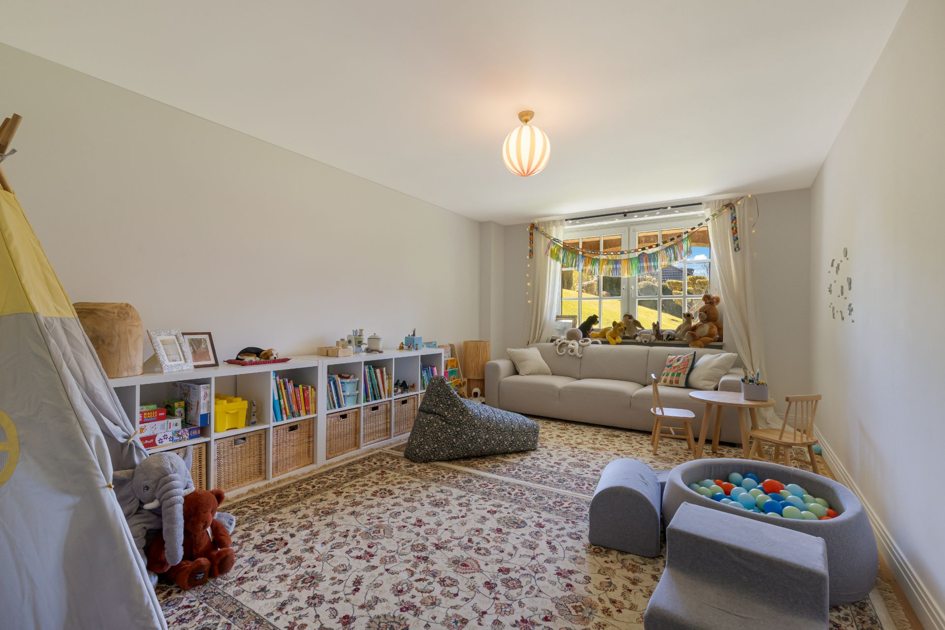 Helles Kinderzimmer mit gemütlichem Sofa, Bücherregal, Spielzeug und großem Teppich.