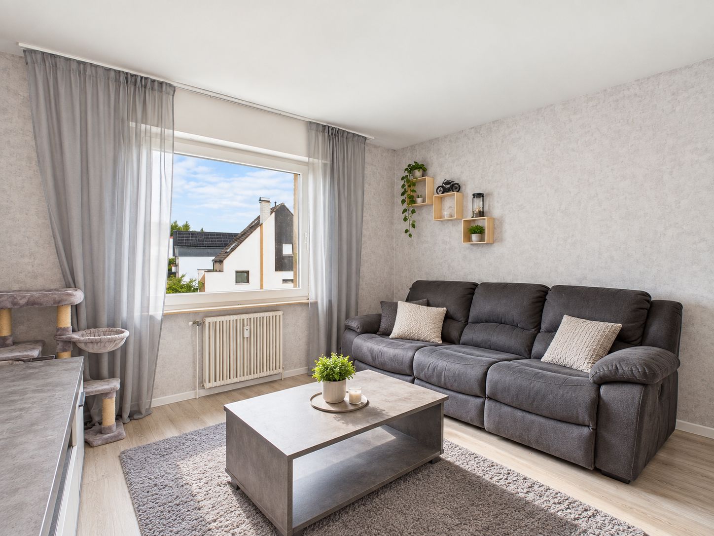 Helles Wohnzimmer mit grauem Sofa, Couchtisch, Teppich und großem Fenster mit Blick nach draußen.