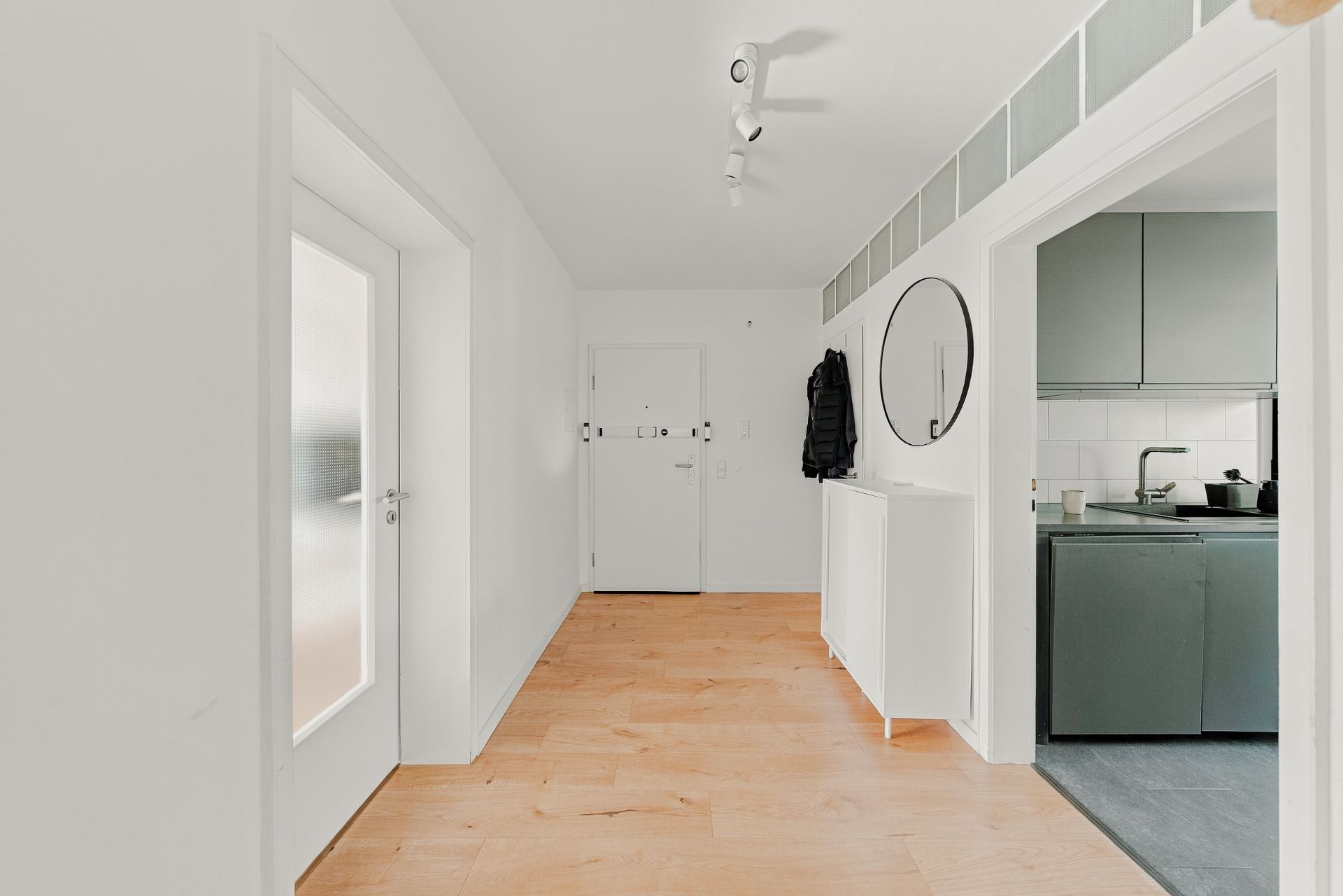 Heller Flur mit Holzfußboden, Garderobe, rundem Spiegel und Blick in eine moderne Küche.