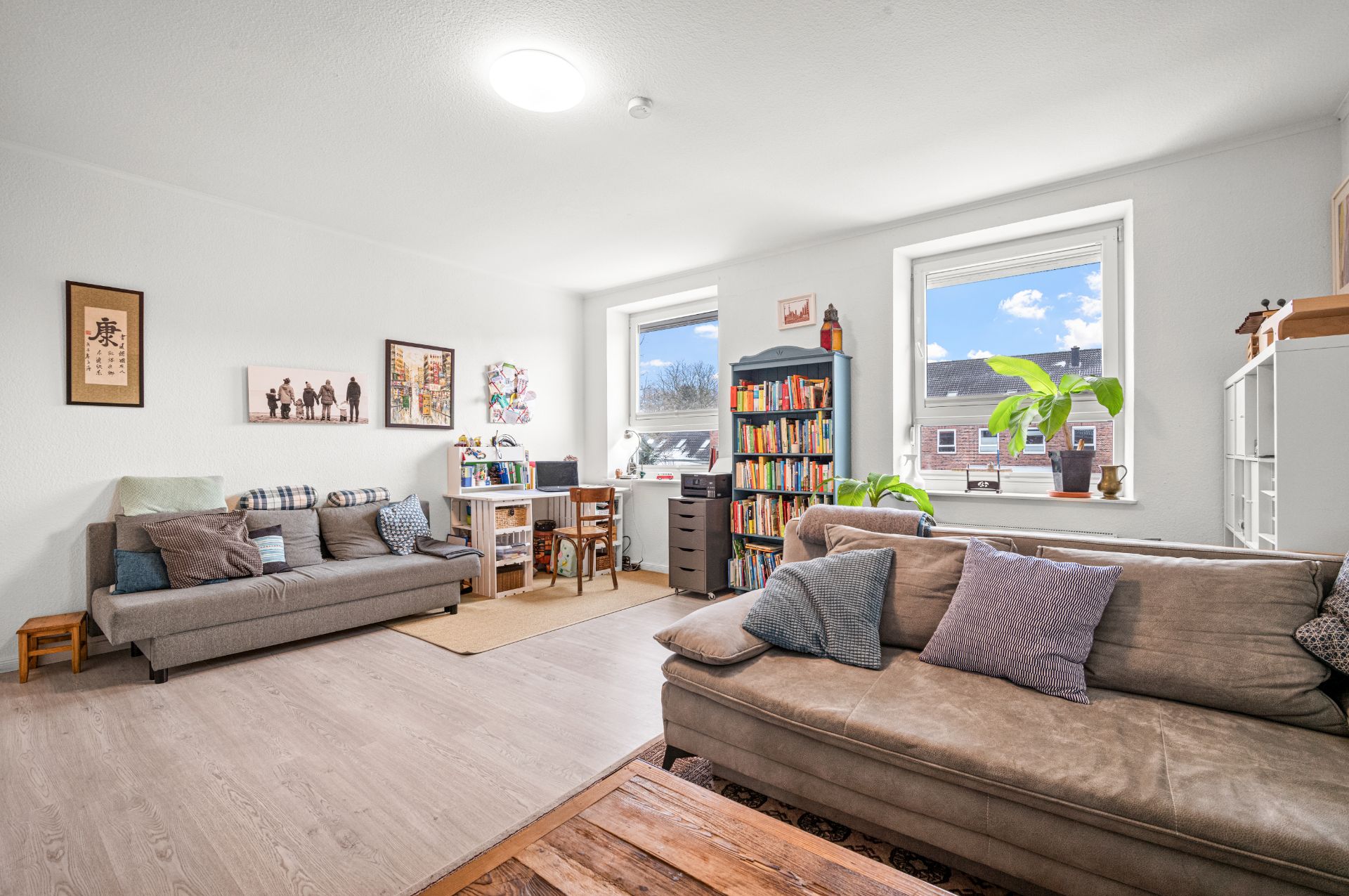 Helles Wohnzimmer mit zwei Sofas, Bücherregal, Schreibtisch und großen Fenstern.