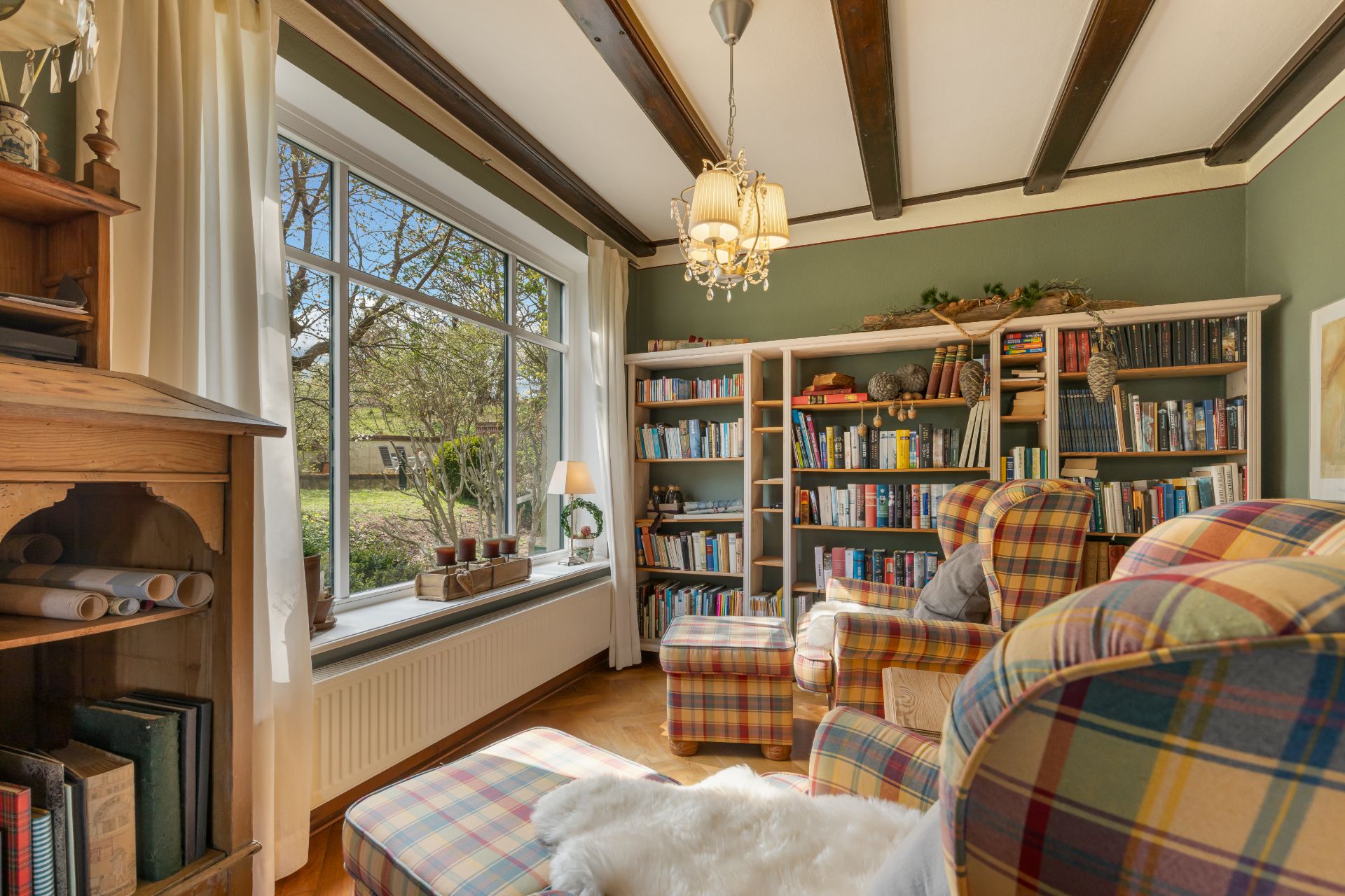 Gemütliches Wohnzimmer mit Sessel, Bücherregalen und großem Fenster mit Blick ins Grüne.