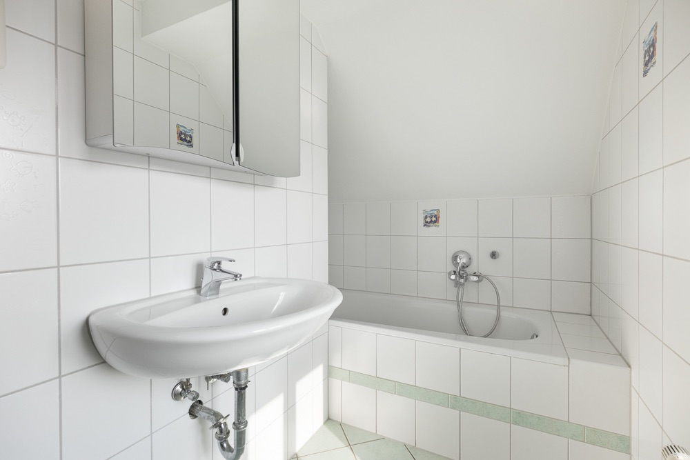 Helles Badezimmer mit weiß geflierten Wänden, Waschbecken, Spiegel und Badewanne.