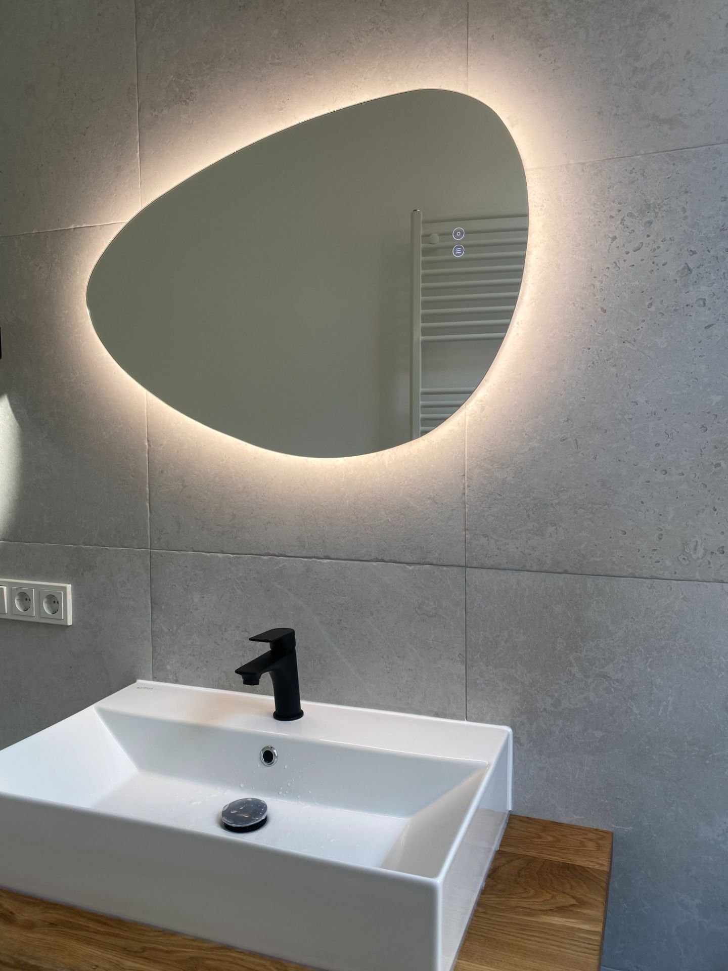Waschbecken mit schwarzem Wasserhahn, Holzablage und beleuchtetem Spiegel im Badezimmer.