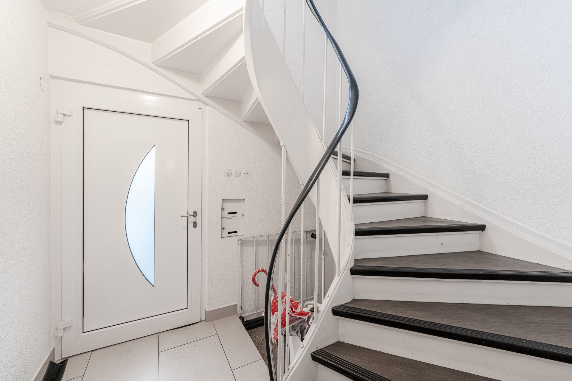 Weiße Haustür und gewendelte Treppe mit schwarzem Handlauf in einem Flur.