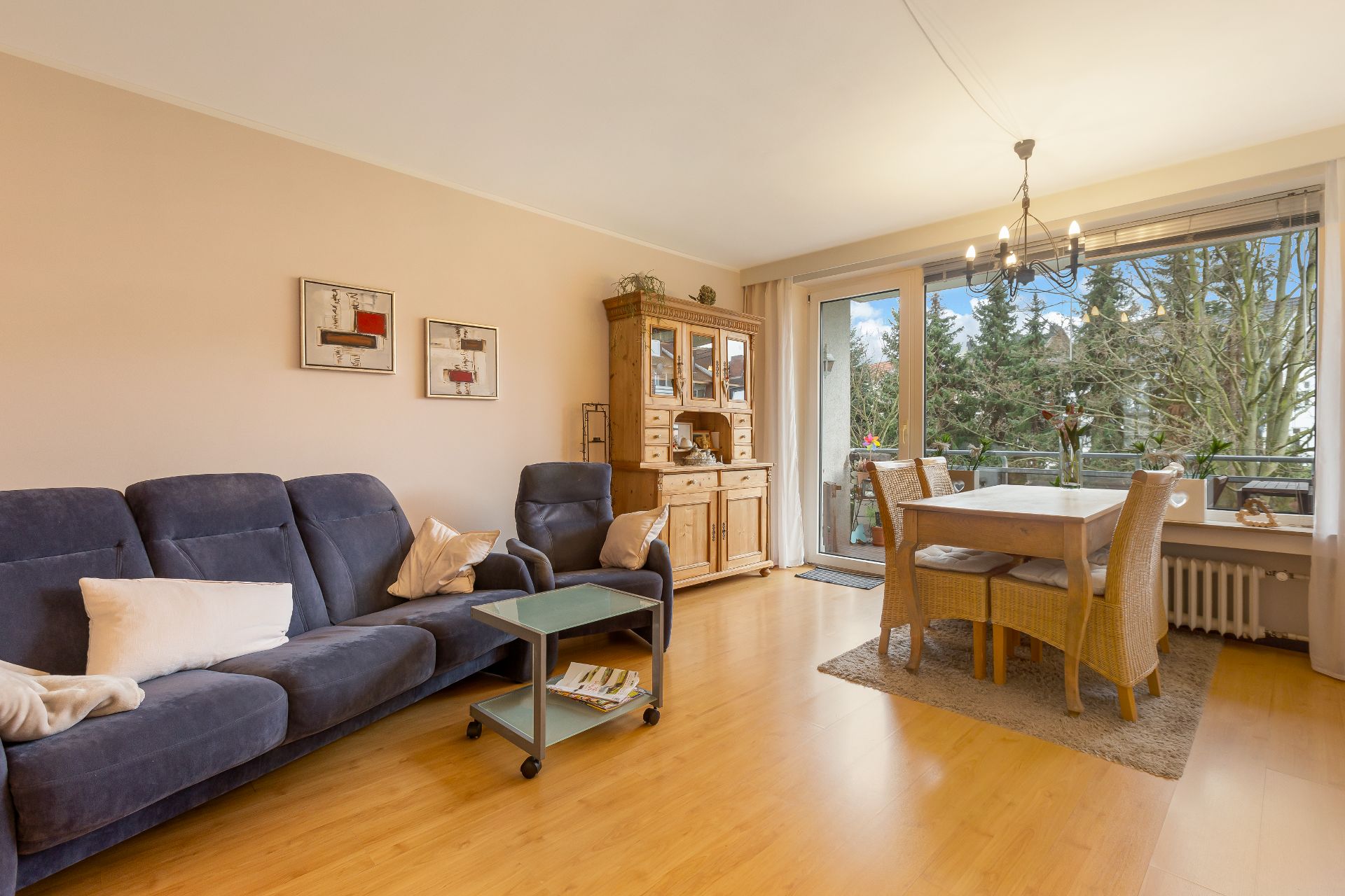 Wohnzimmer mit blauer Couch, Esstisch aus Holz, Kommode und großem Fenster zum Balkon.