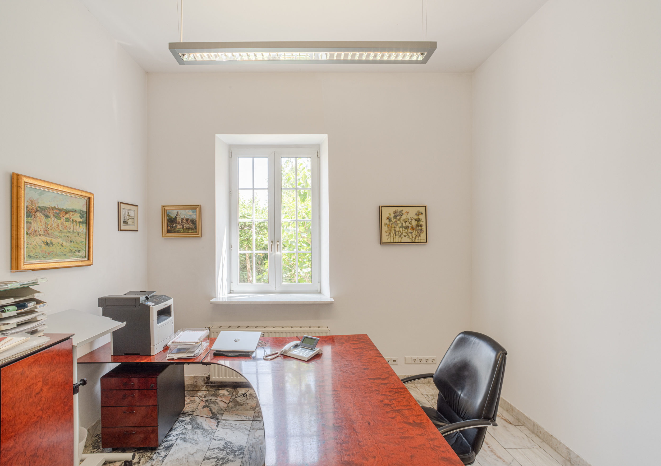 Helles Arbeitszimmer mit rotem Schreibtisch, schwarzem Stuhl und Fenster, umgeben von Gemälden an weißen Wänden.