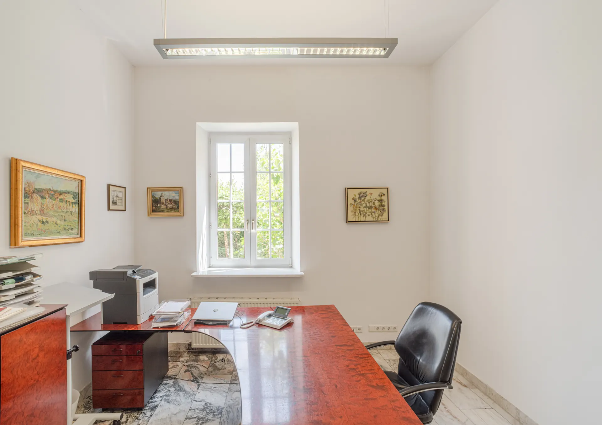 Helles Arbeitszimmer mit rotem Schreibtisch, schwarzem Stuhl und Fenster, umgeben von Gemälden an weißen Wänden.