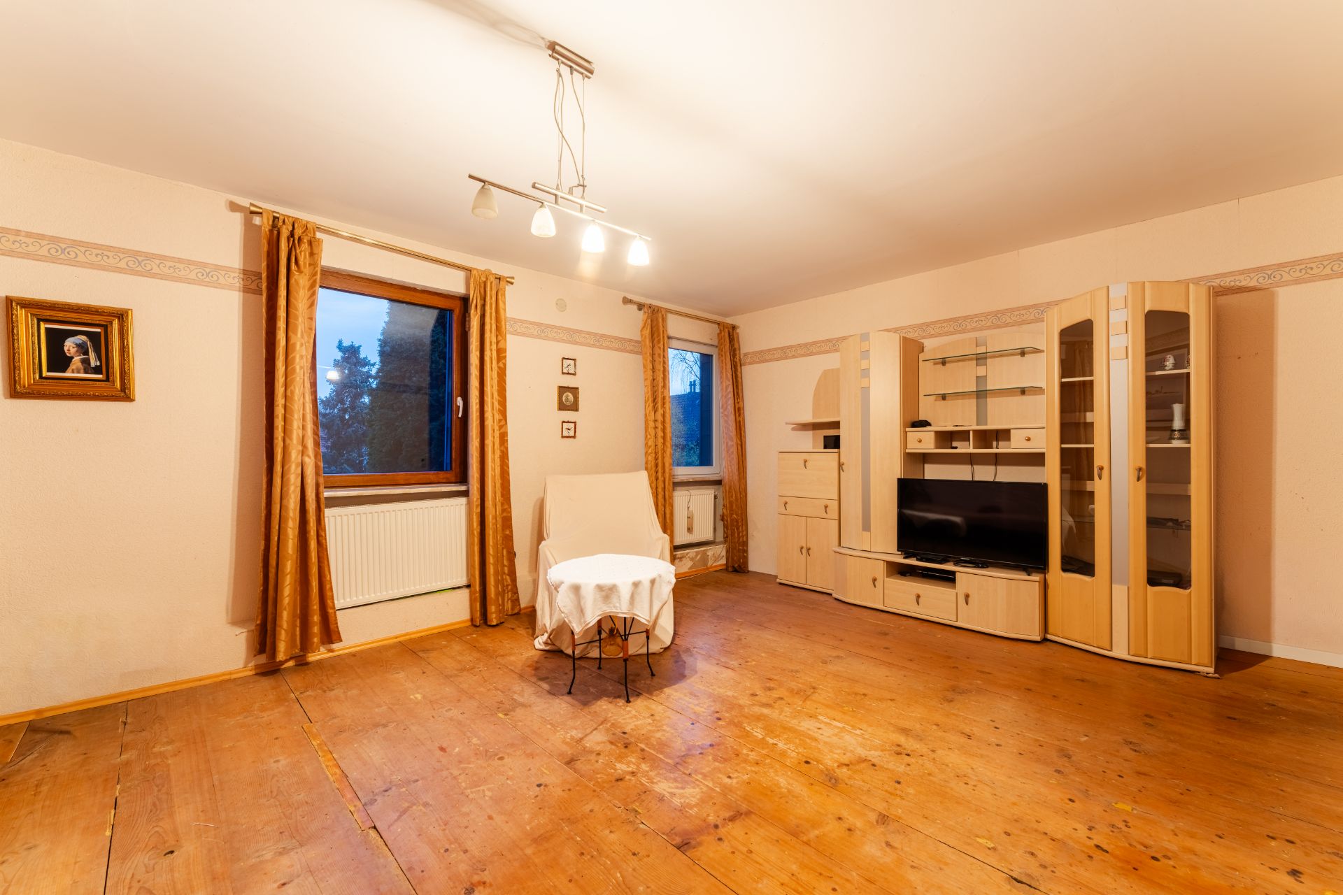Wohnzimmer mit Holzfußboden, Regalwand, Sessel, kleinem Tisch und zwei Fenstern mit Vorhängen.