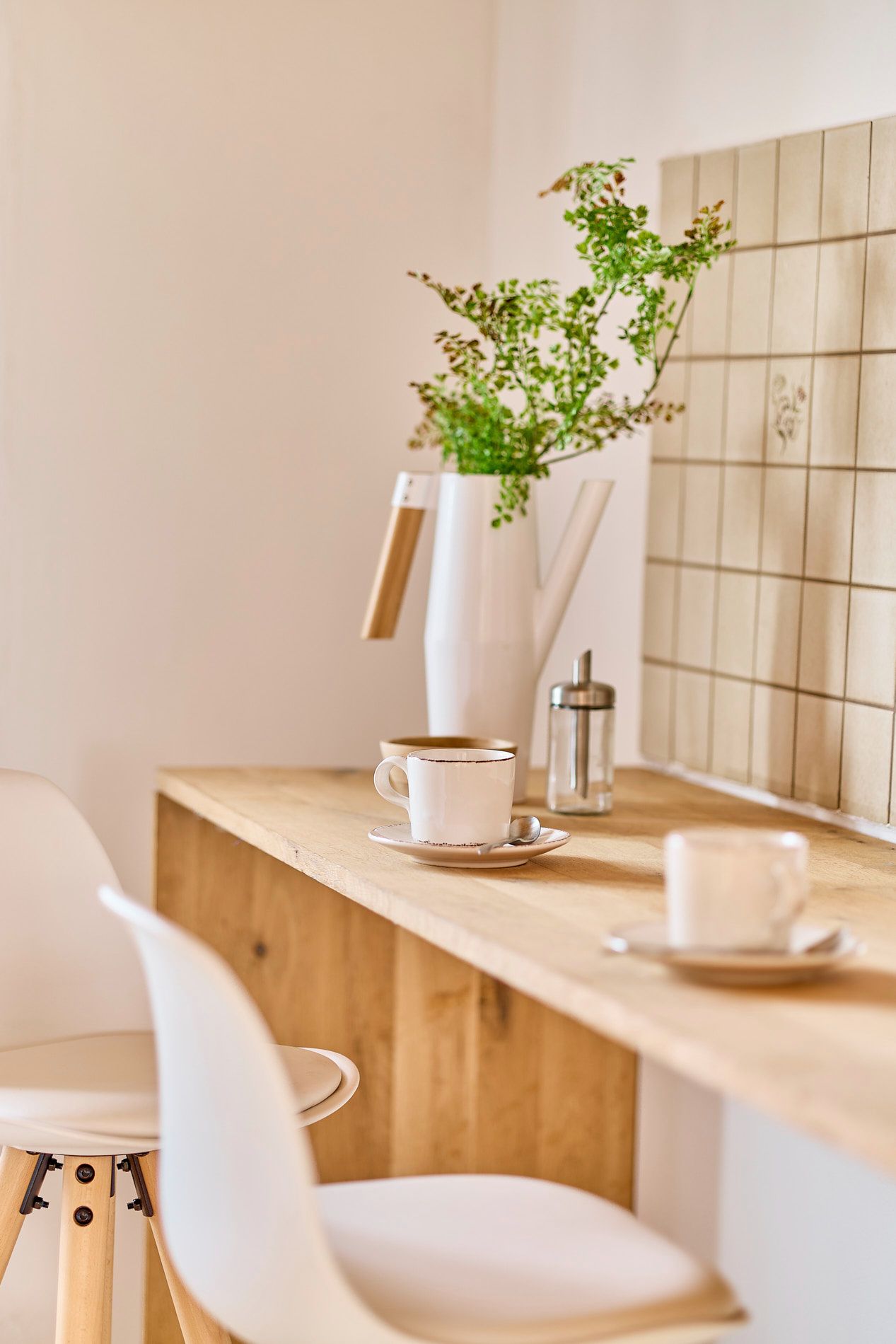 Holztheke mit zwei weißen Stühlen, Tassen, Zuckerdose und Vase mit Zweigen vor Fliesenwand.
