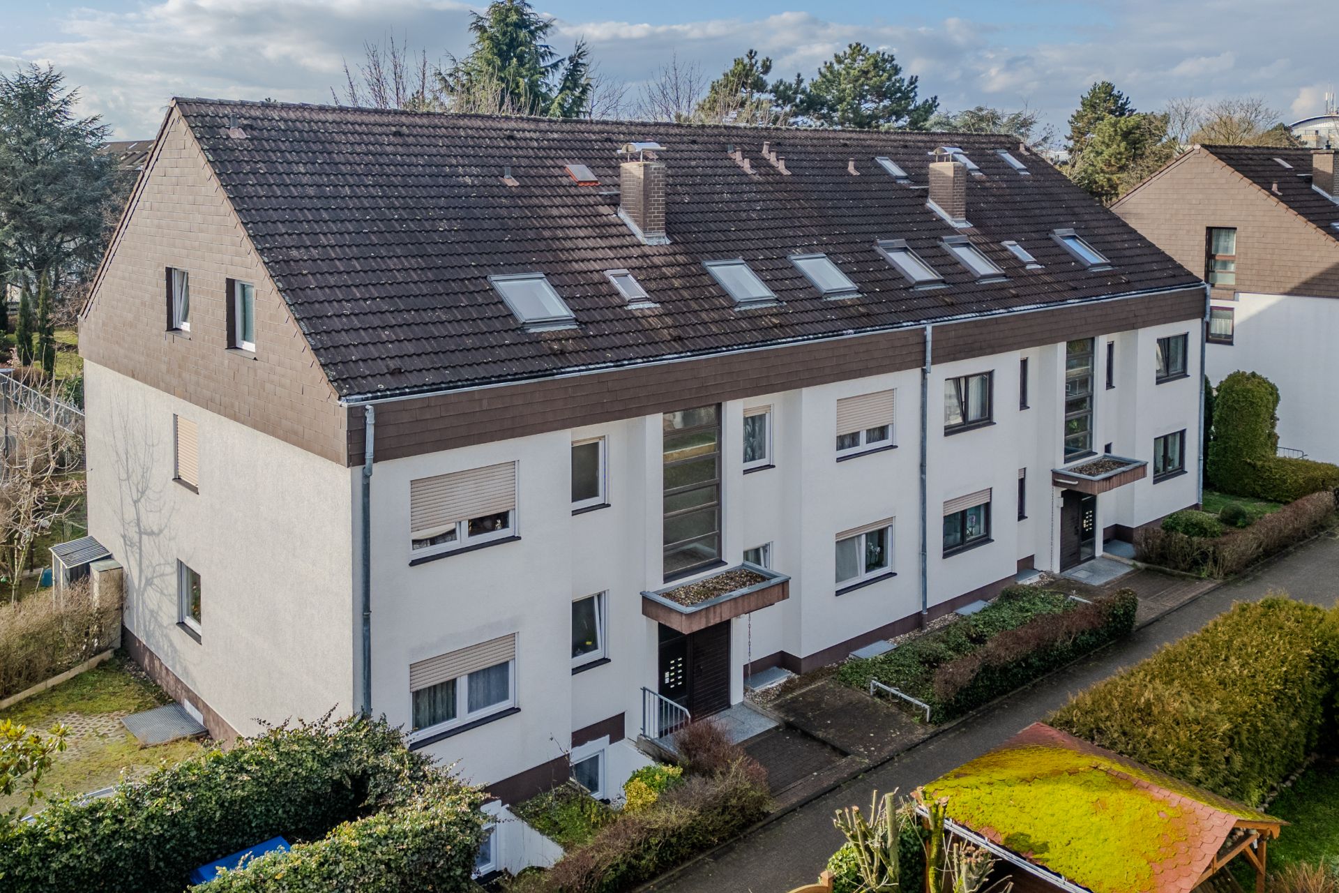 Mehrfamilienhaus mit braunem Satteldach, weißen Fassaden und mehreren Eingängen, umgeben von Büschen.