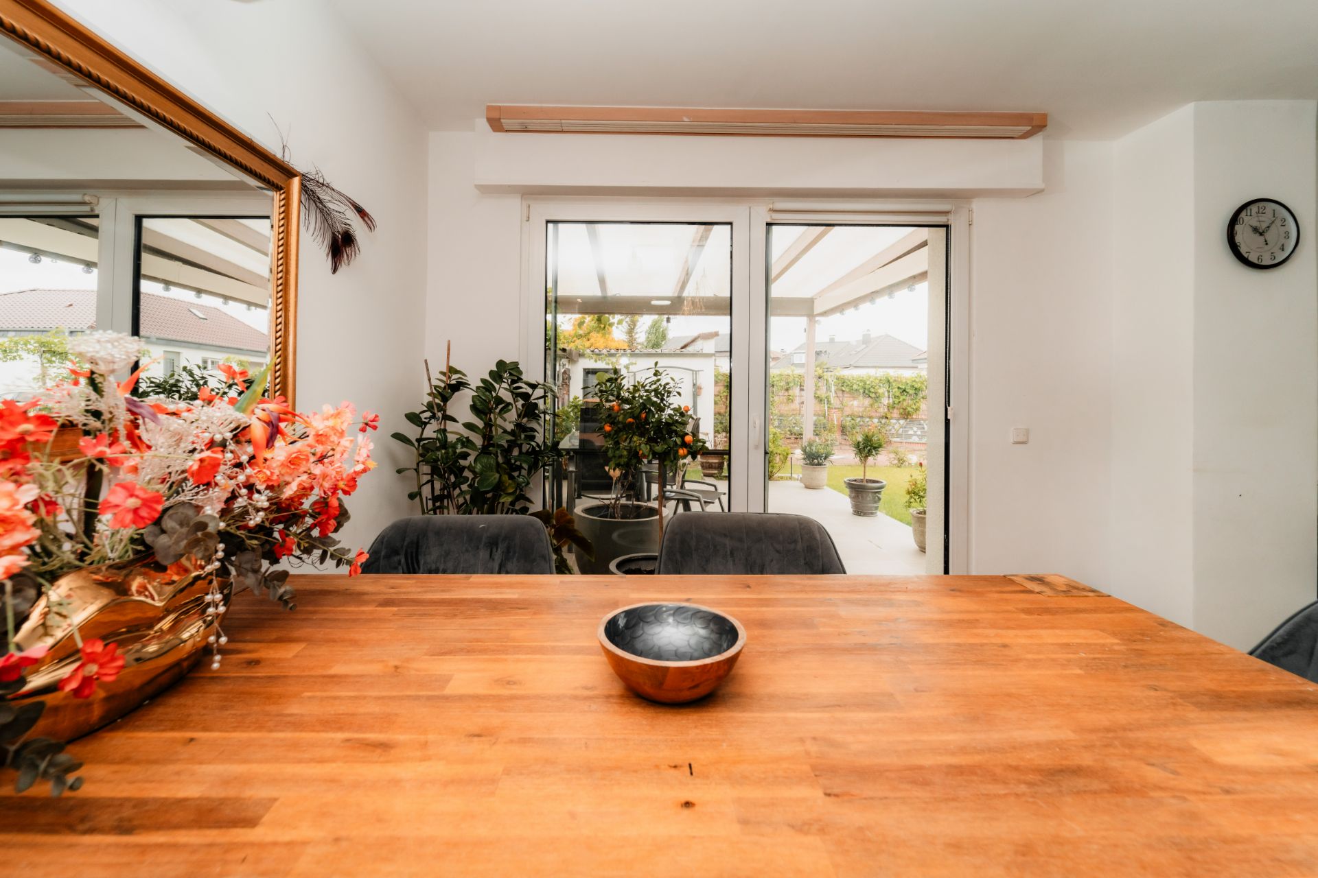 Esszimmer mit Holztisch, Schale, Blumen, Spiegel und Blick auf Terrasse mit Pflanzen.