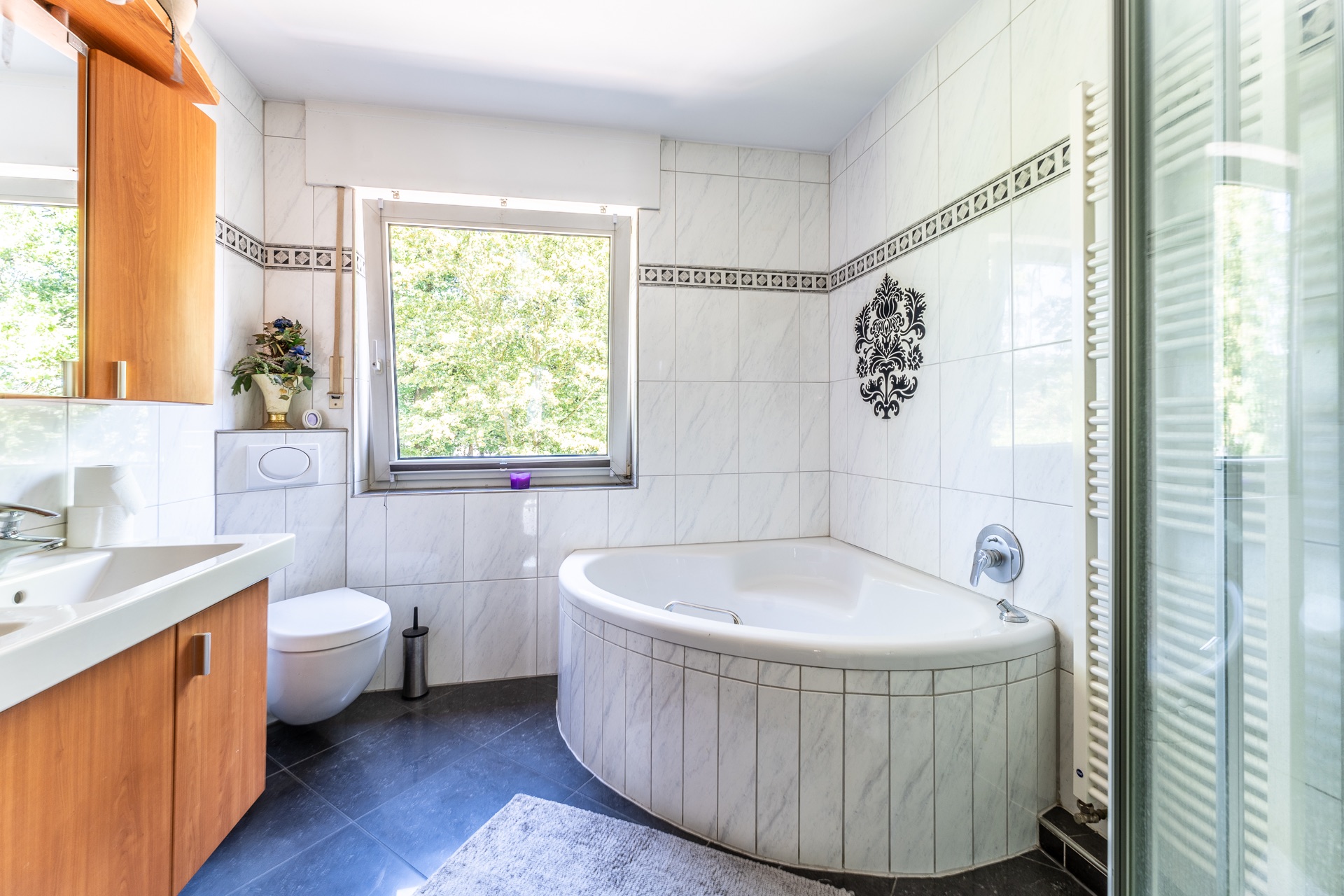 Helles Badezimmer mit Eckbadewanne, Waschbecken, WC und Fenster mit Blick ins Grüne.
