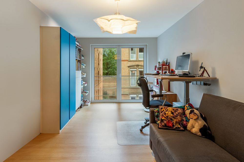 Helles Zimmer mit Sofa, Schreibtisch, Computer, blauem Schrank und Fenster mit Blick nach draußen.