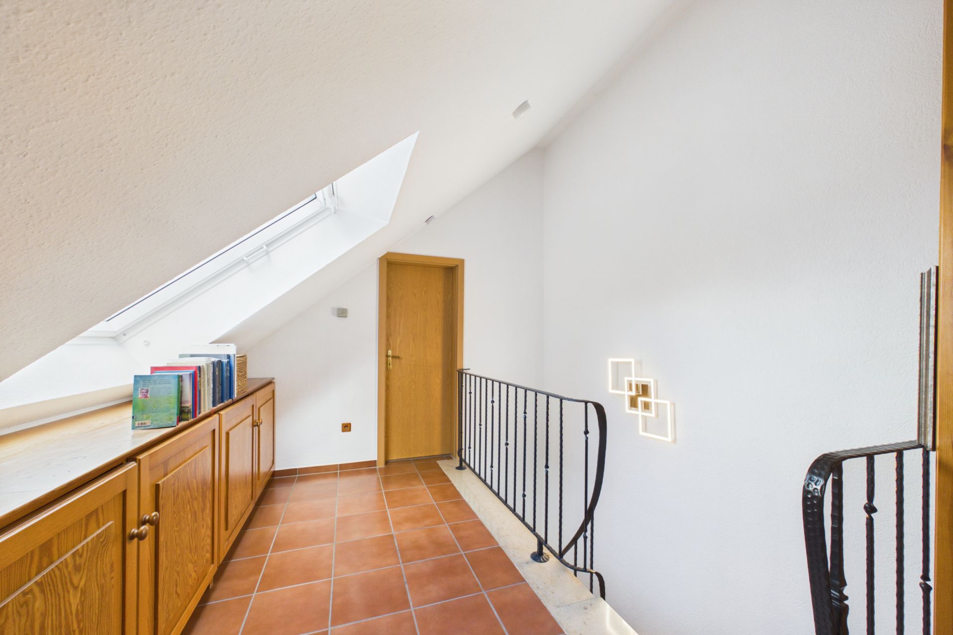 Heller Flur mit Dachschräge, Terrakottafliesen, Holzschränken und Geländer, Bücher auf dem Schrank.