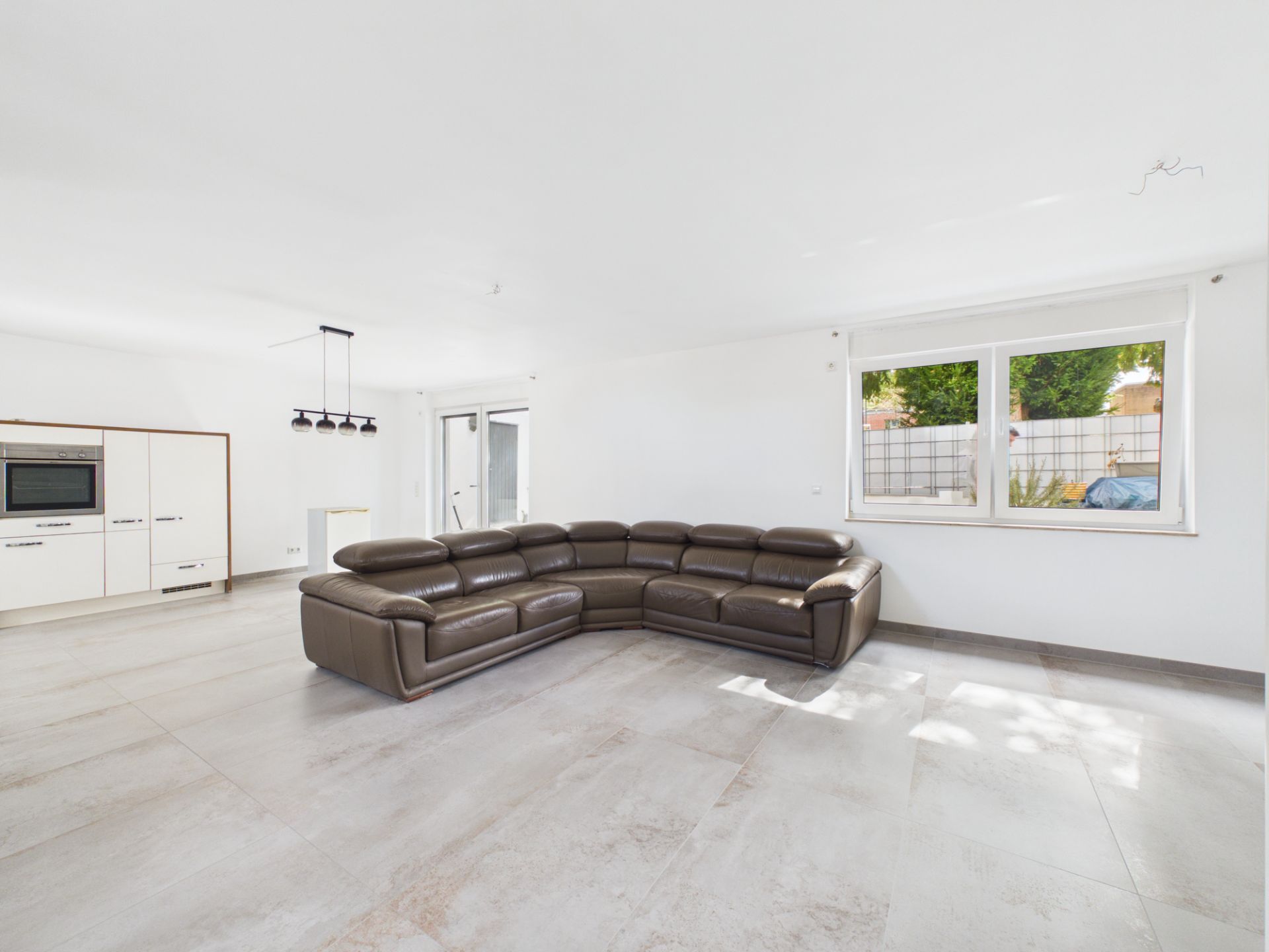 Helles Wohnzimmer mit braunem Ecksofa, großem Fenster und offener Einbauküche.