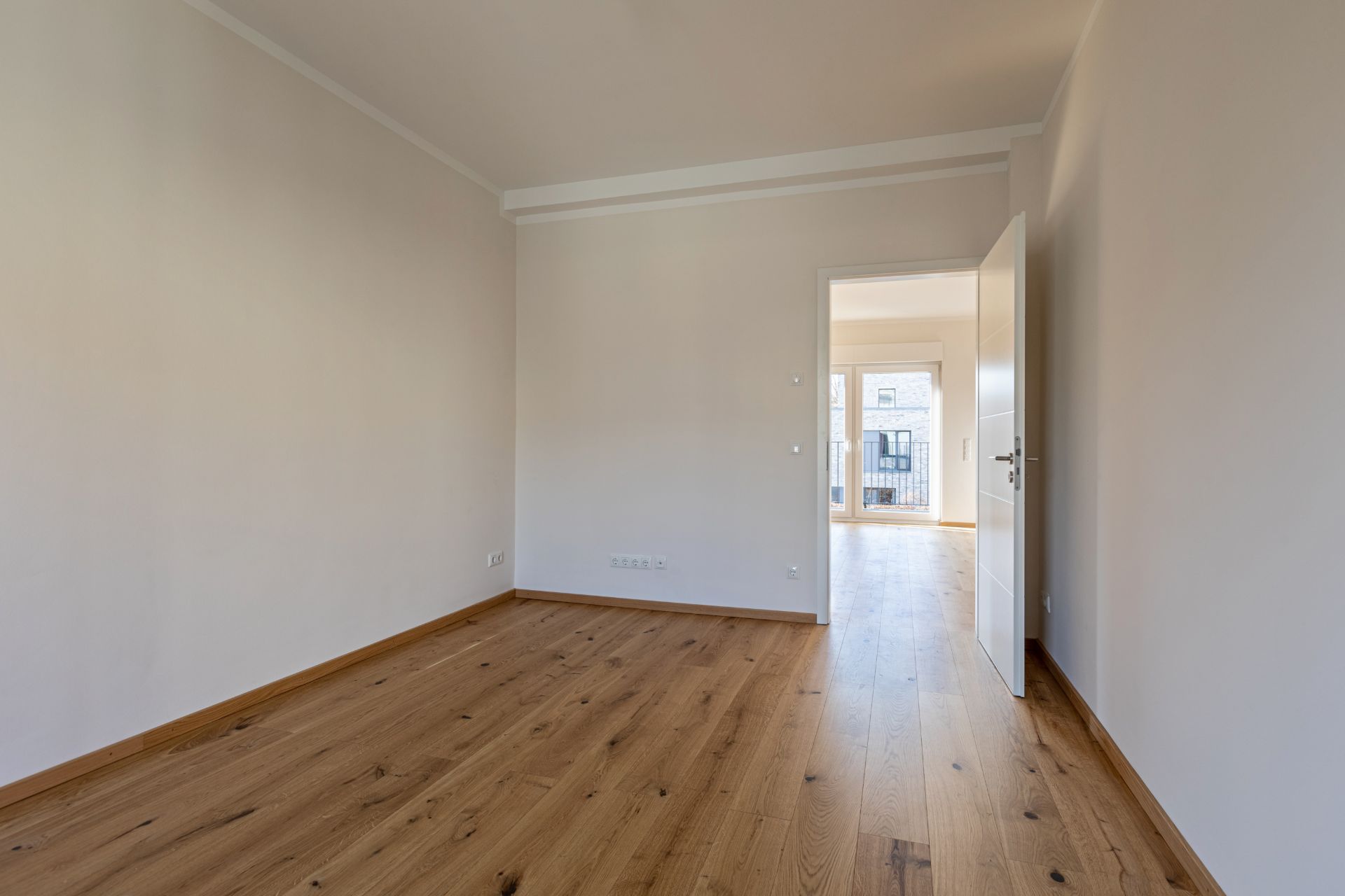 Leerer Raum mit Holzboden, weißen Wänden und geöffneter Tür zu einem hellen Zimmer.