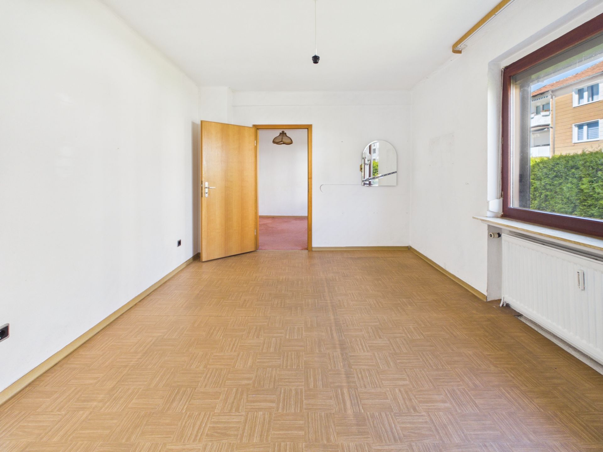 Leerer Raum mit braunem Boden, großem Fenster, offen stehender Holztür und Spiegel an der Wand.