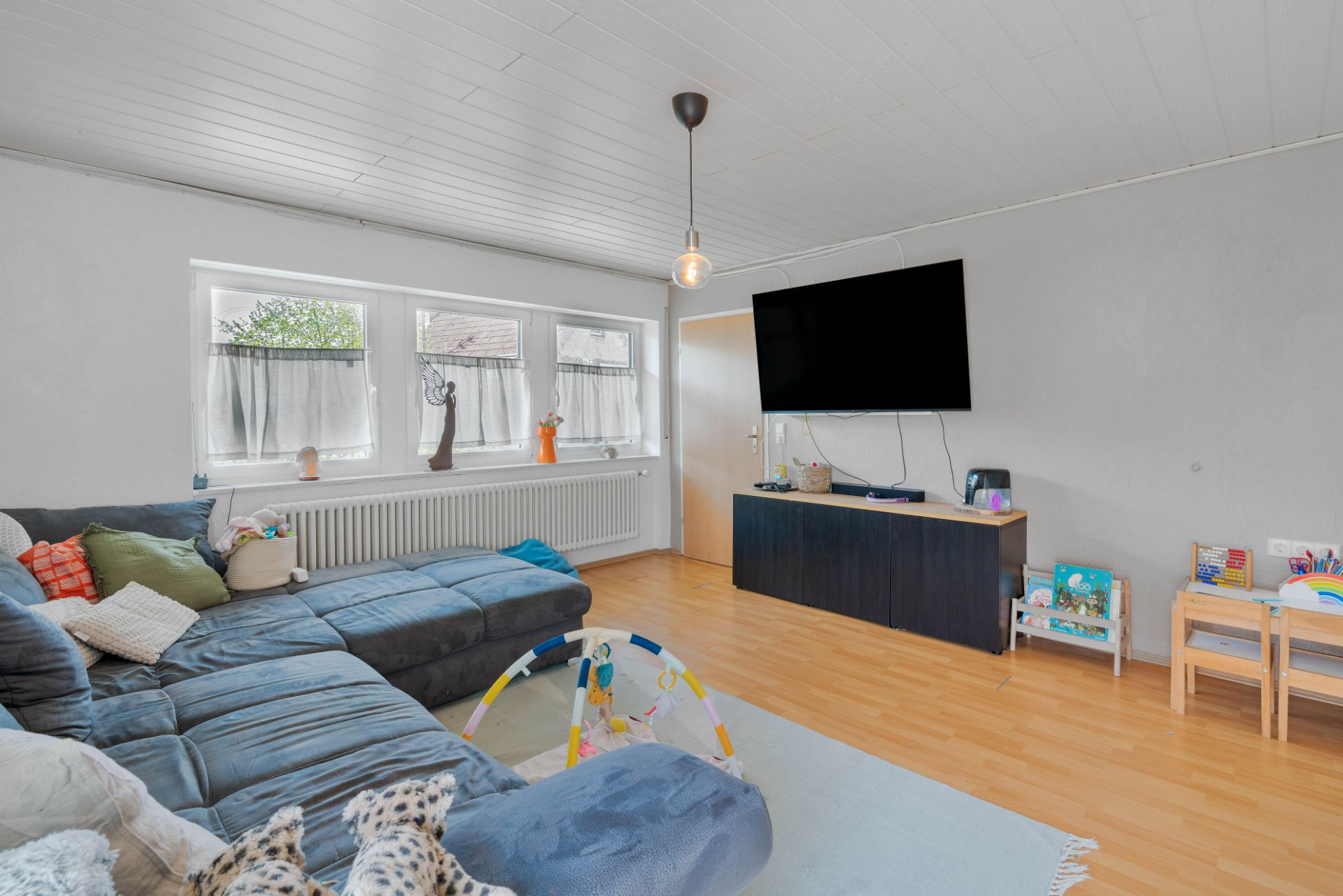 Wohnzimmer mit großem Sofa, Fernseher an der Wand, Spielzeug und drei Fenstern.