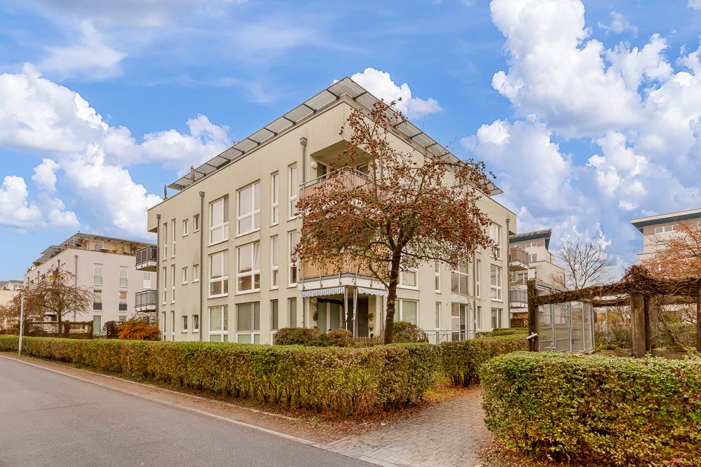 Modernes Wohngebäude mit Balkonen, umgeben von Hecken und Bäumen an einer ruhigen Straße.
