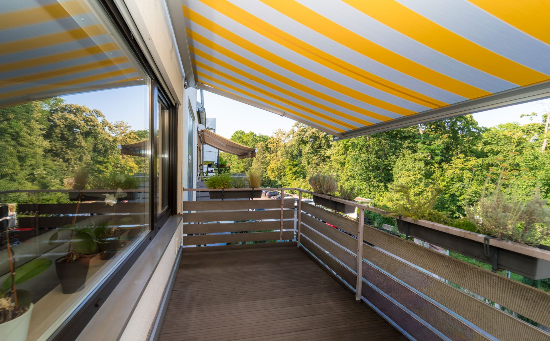 Balkon mit Holzgeländer, gelb-weiß gestreifter Markise und Pflanzen, Blick ins Grüne.