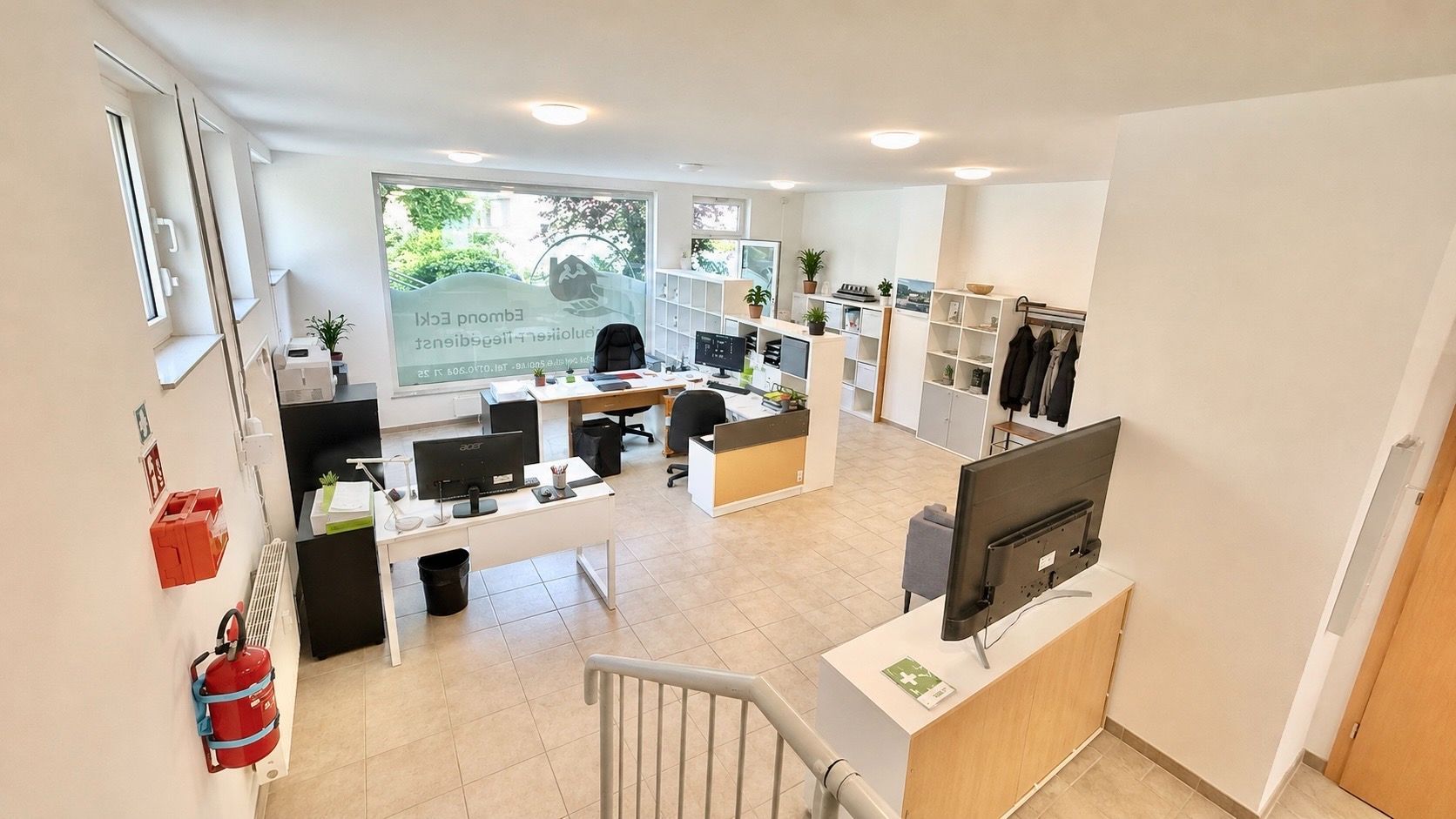 Heller Büroraum mit mehreren Schreibtischen, Computern und Regalen, aufgenommen von einer Treppe.