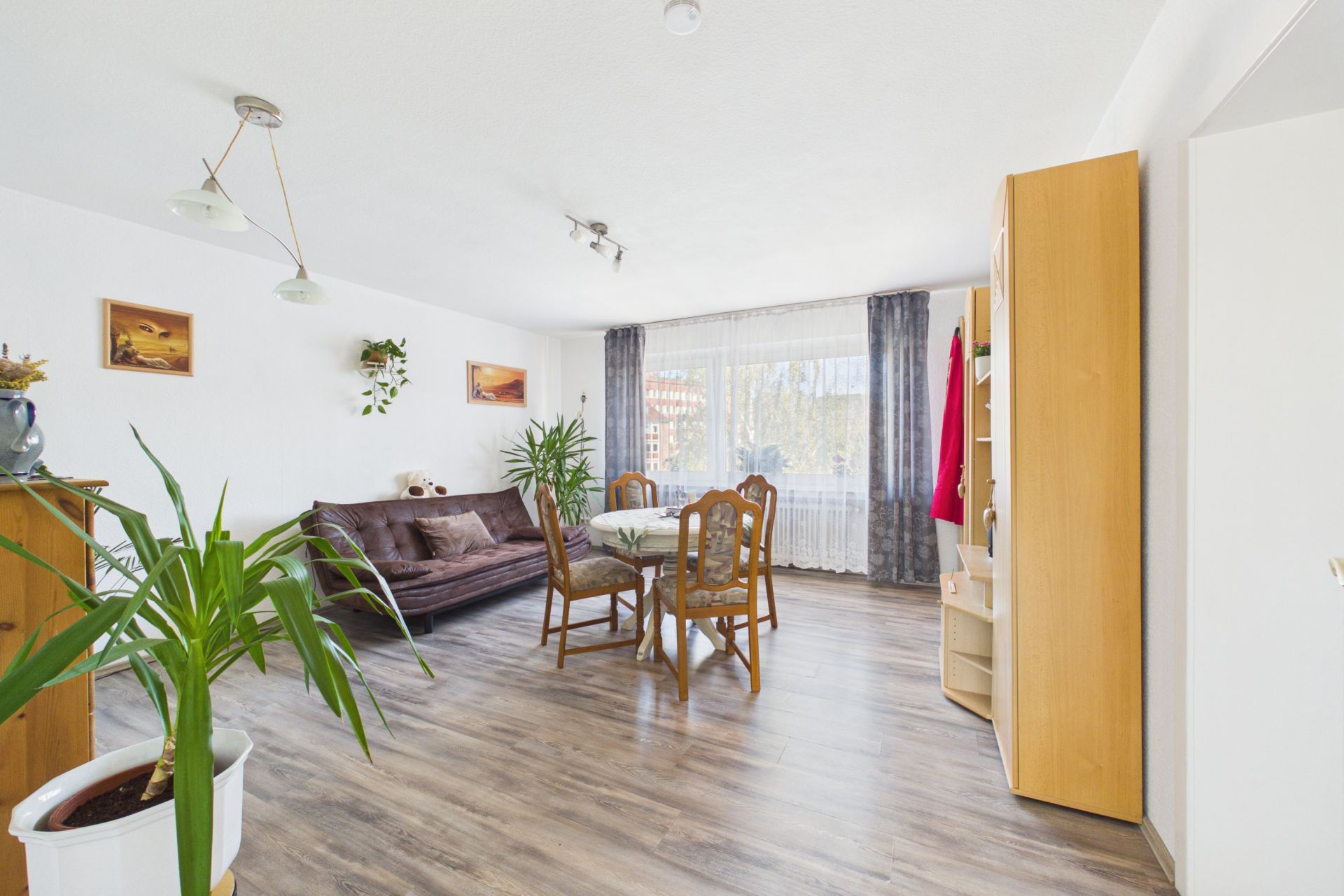 Helles Wohnzimmer mit braunem Sofa, Esstisch, Stühlen, Pflanzen und großem Fenster.