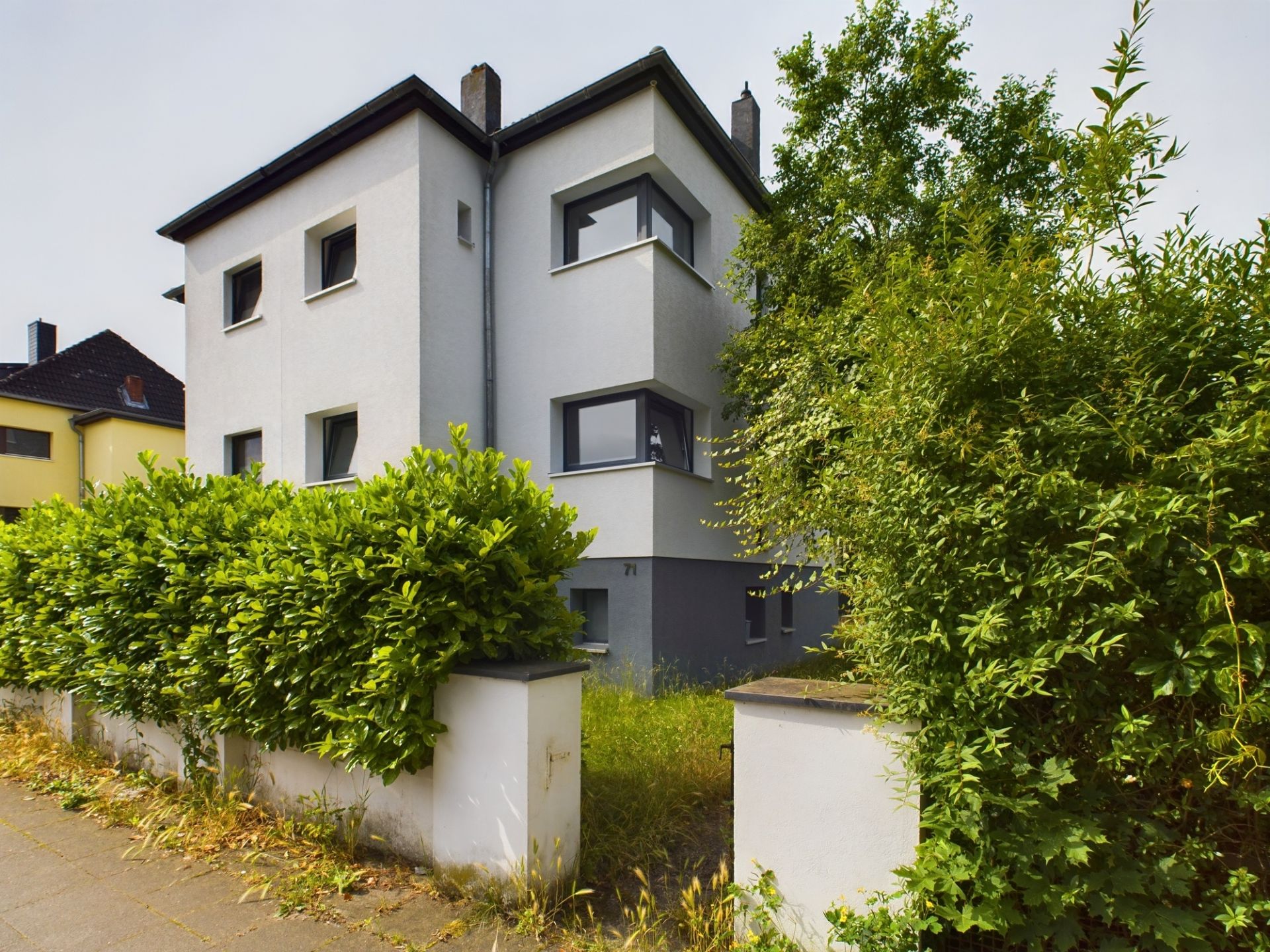 Modernes Wohnhaus mit weißer Fassade, großen Fenstern und grüner Hecke vor dem Haus.