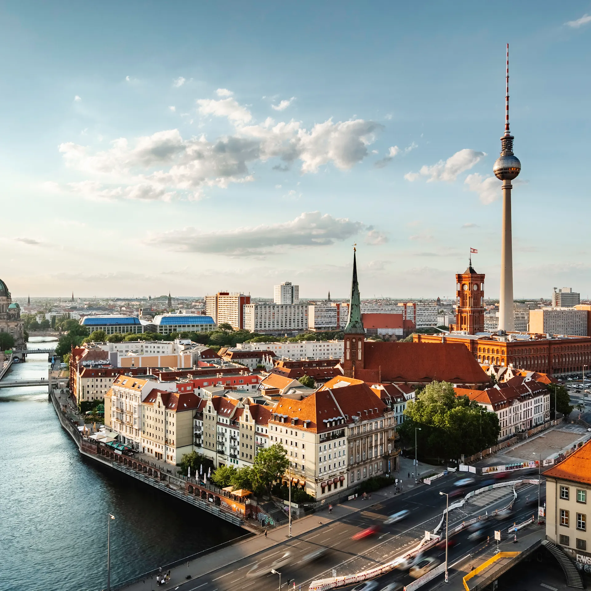 Stadtansicht mit Fluss, roten Dächern und Fernsehturm unter blauem Himmel.