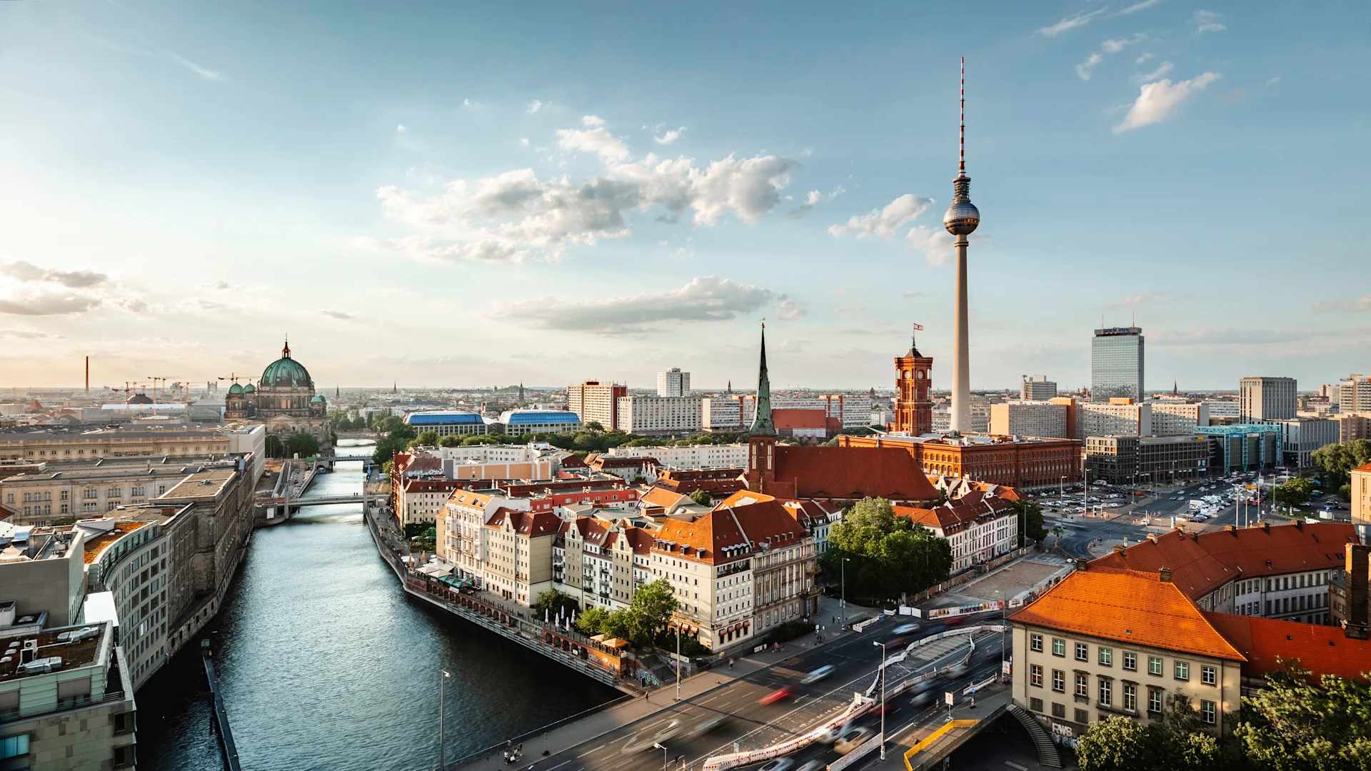 Stadtansicht mit Fluss, roten Dächern und Fernsehturm unter blauem Himmel.
