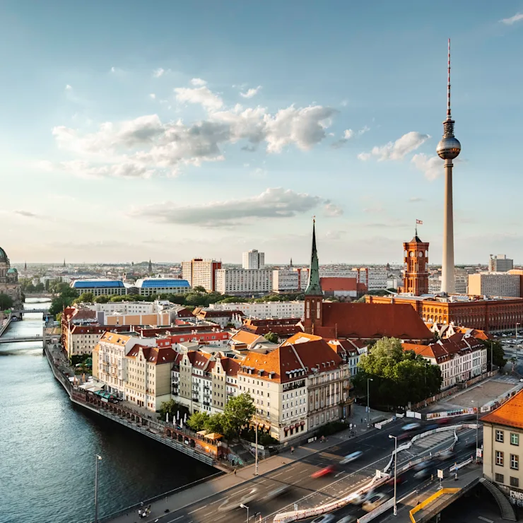 Stadtansicht mit Fluss, roten Dächern und Fernsehturm unter blauem Himmel.