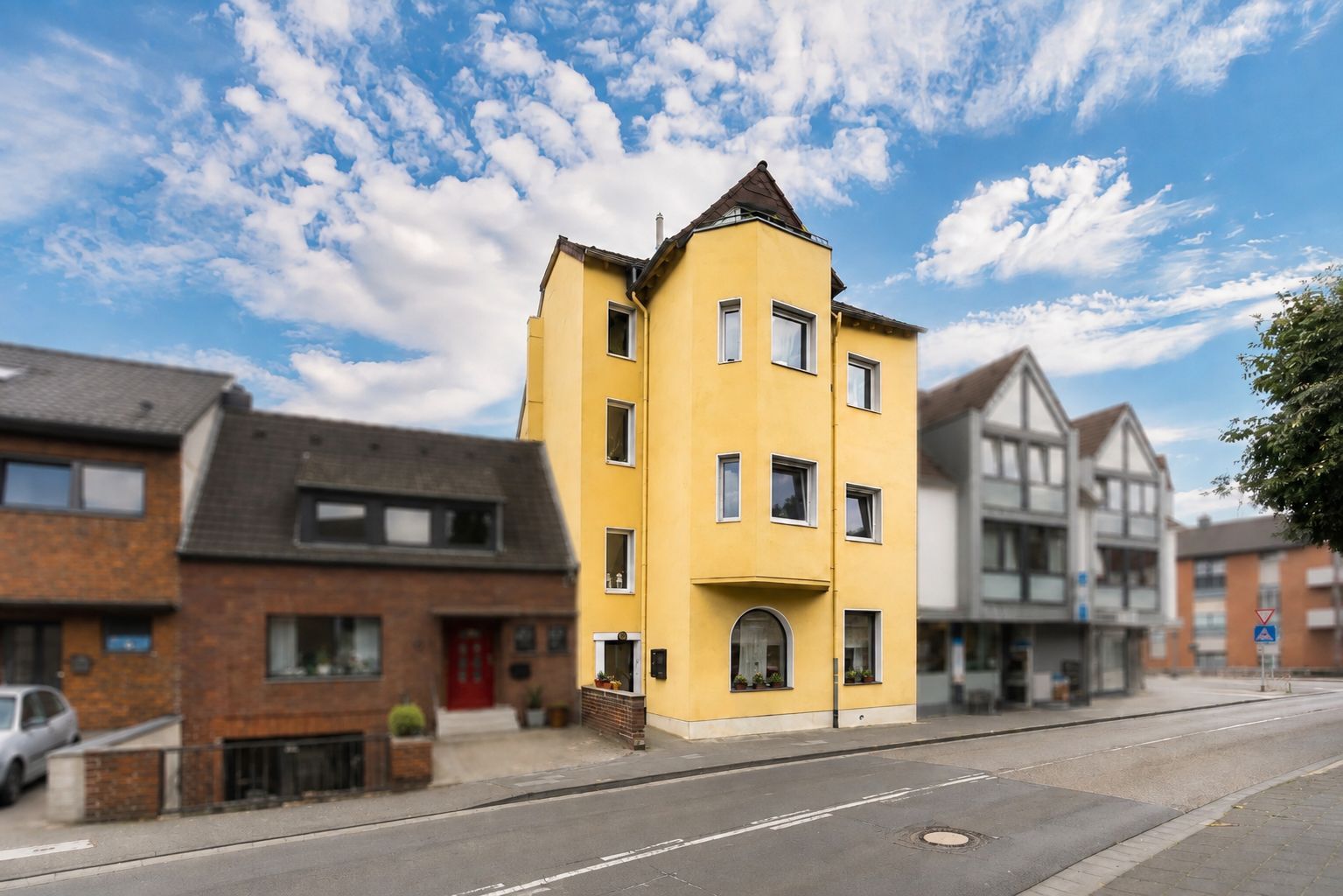 Gelbes Wohnhaus mit vier Etagen und spitzem Dach, an einer Straße gelegen.