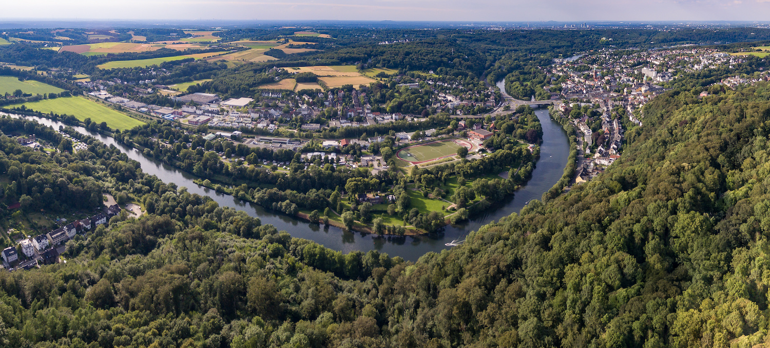 Luftaufnahme einer Wohnsiedlung mit Fluss, Feldern und viel Wald im Vordergrund.