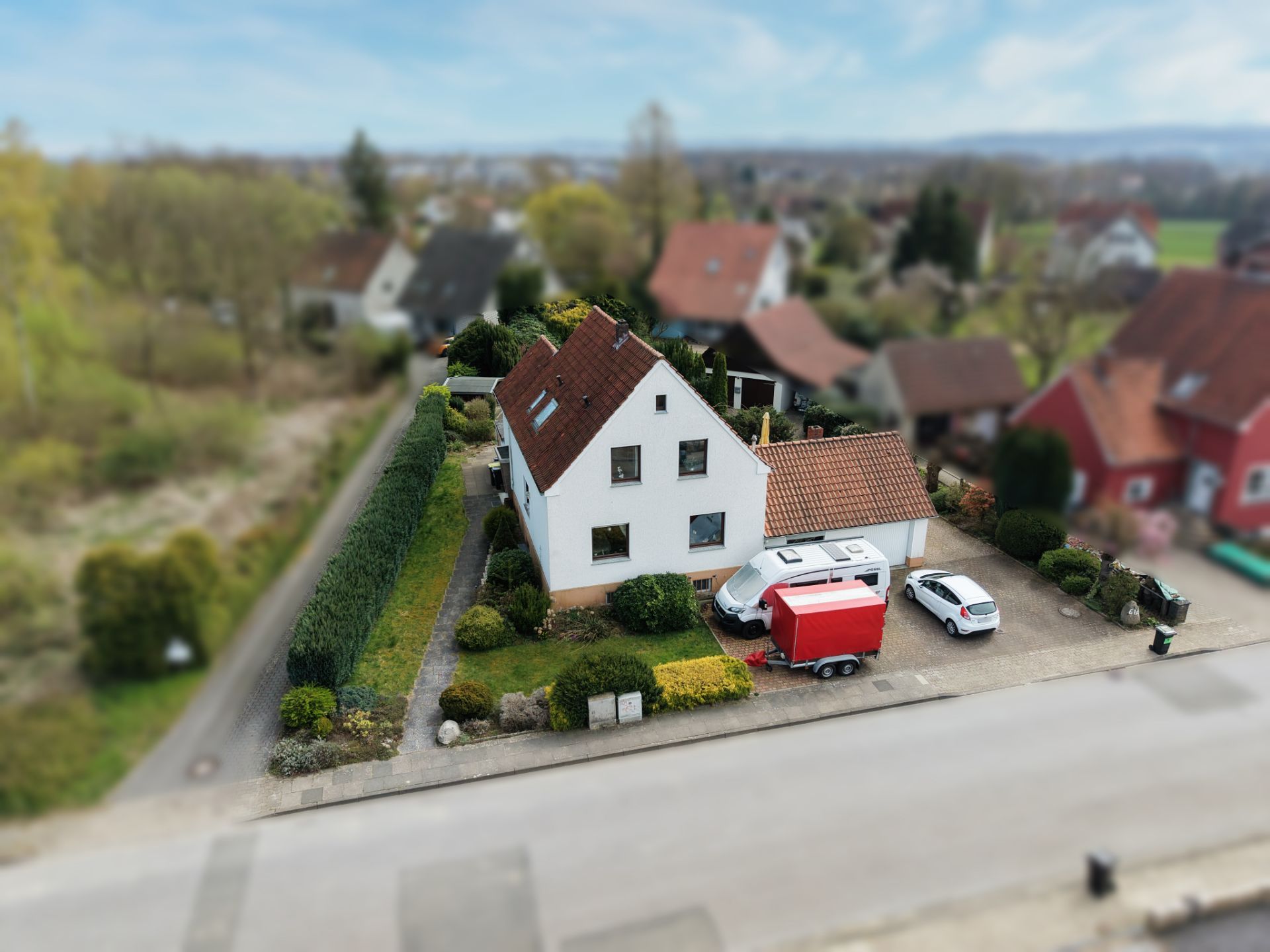Weißes Einfamilienhaus mit rotem Dach, Einfahrt, zwei Autos und Garten, aus der Vogelperspektive.