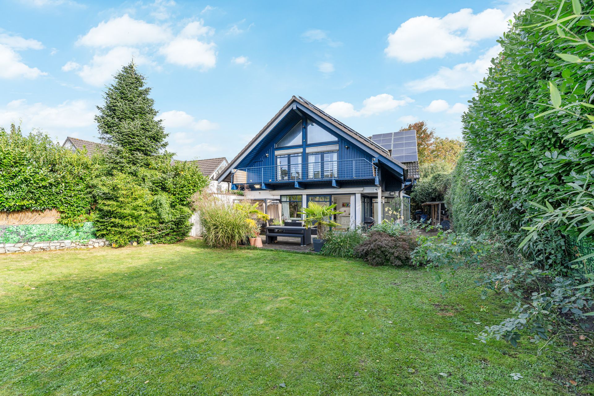 Modernes Haus mit Balkon, großer Glasfront und Garten, umgeben von Hecken und Pflanzen.