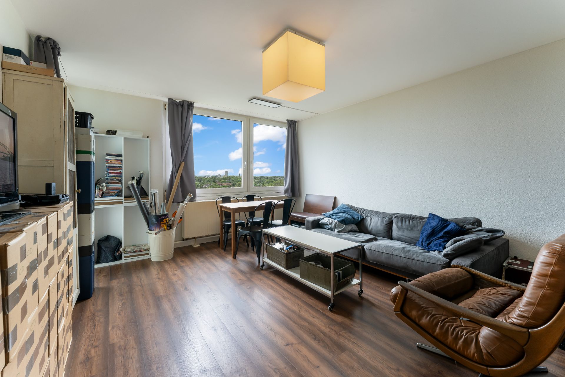 Helles Wohnzimmer mit Sofa, Sessel, Couchtisch, Esstisch und Regal vor einem großen Fenster.