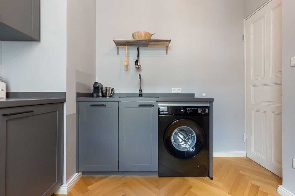 Kompakte Küche mit grauen Schränken, Waschmaschine, Spüle und Regal mit Küchenutensilien.