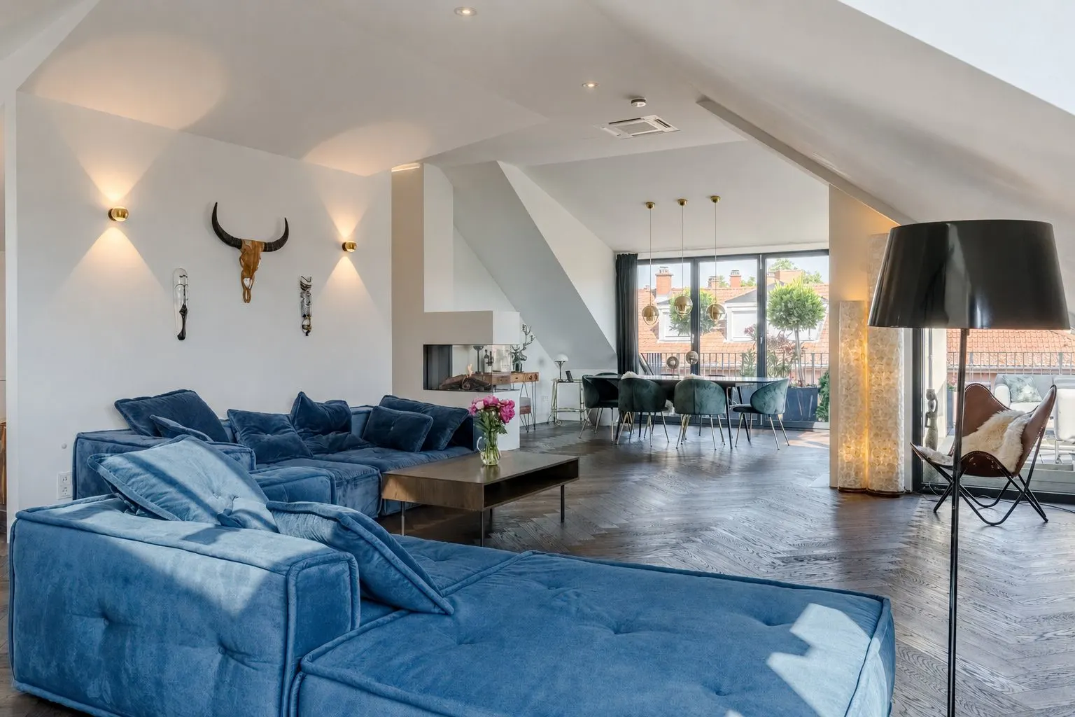 Modernes Wohnzimmer mit blauen Sofas, Kamin, Esstisch und großen Fenstern.