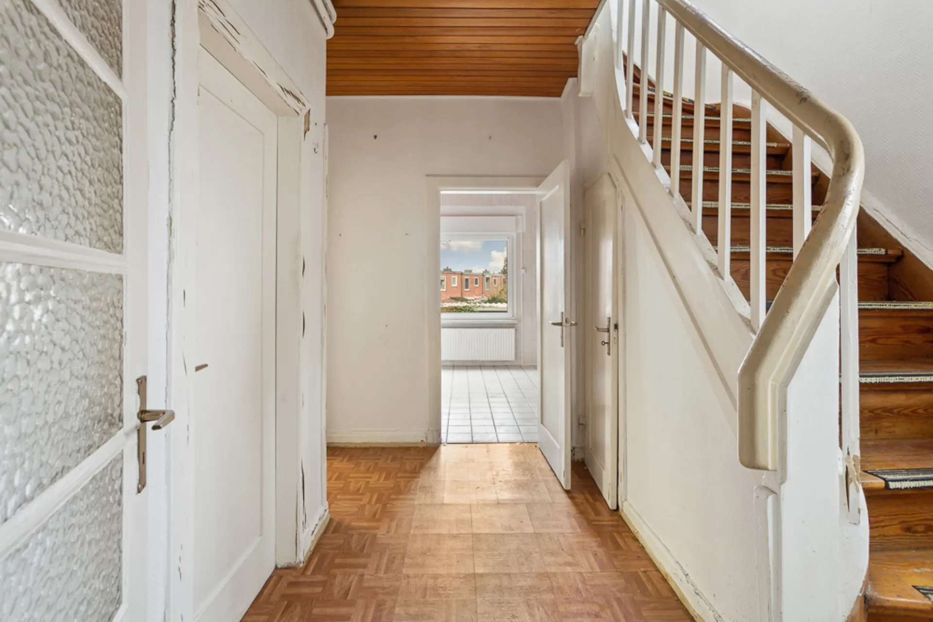 Flur mit Holztreppe, Türen und Blick in einen hellen Raum mit Fenster.