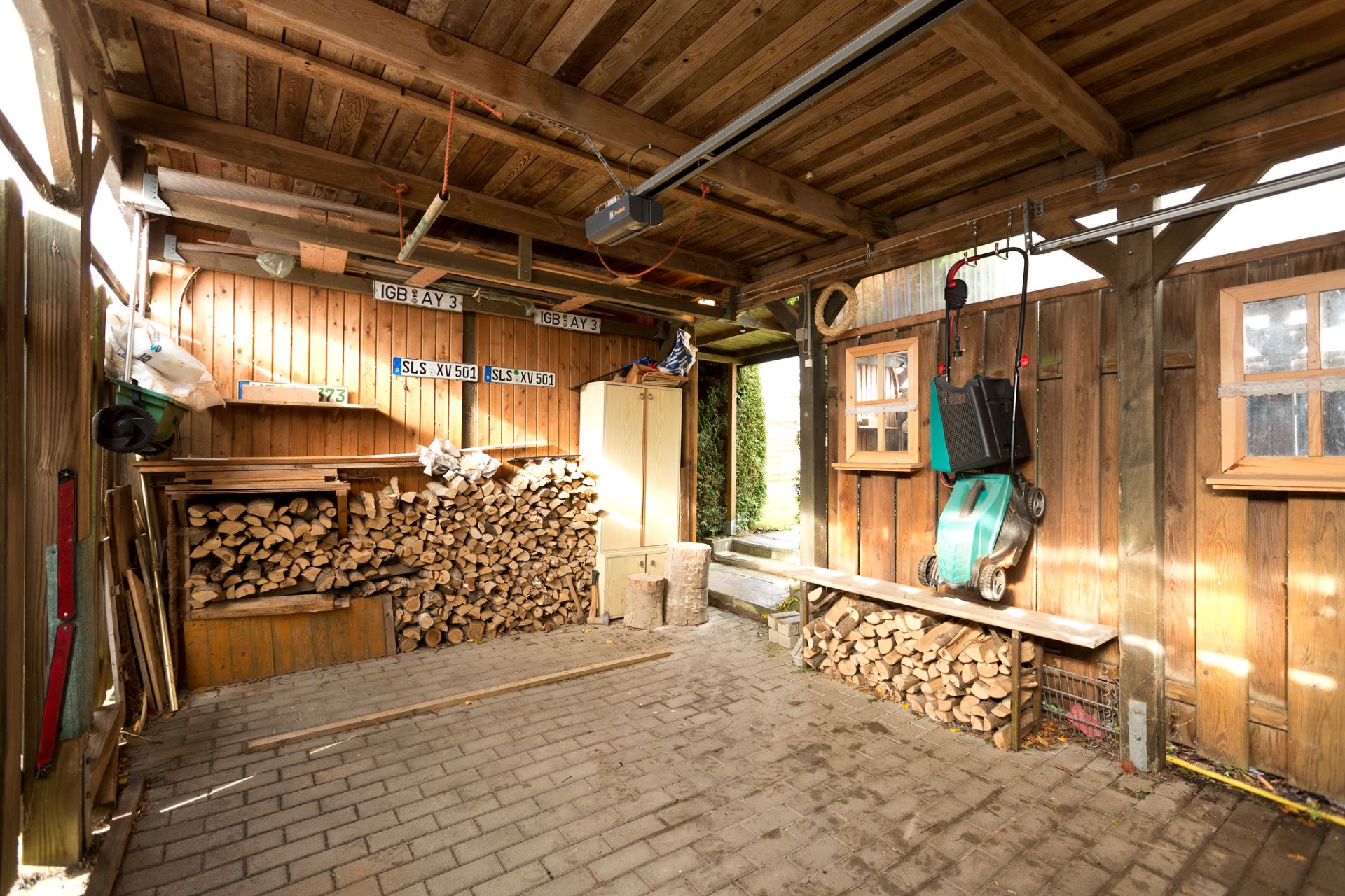 Überdachte Holzhütte mit gestapeltem Brennholz, Gartengeräten und gepflastertem Boden.