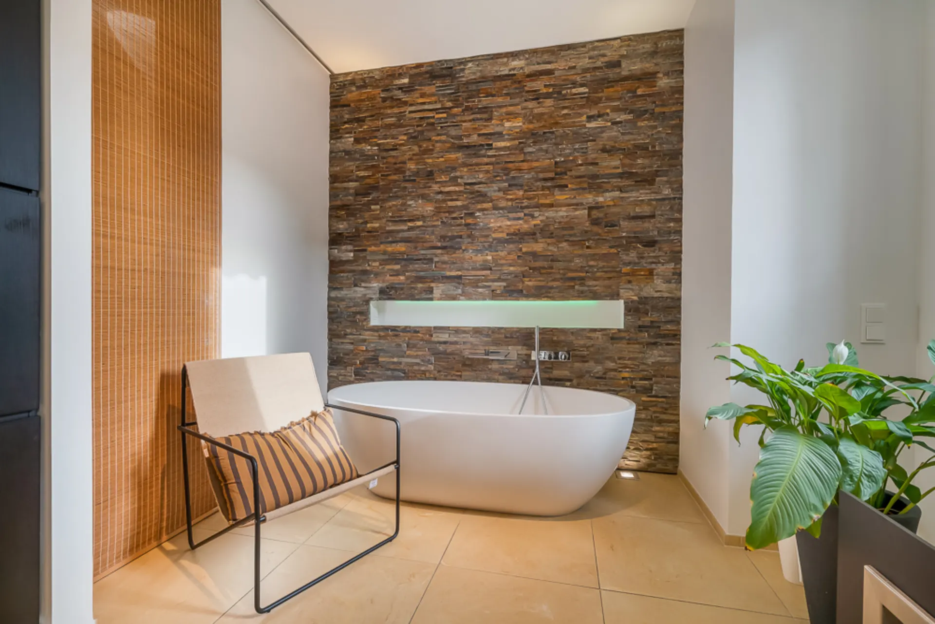 Modernes Badezimmer mit freistehender Badewanne, Stuhl, grüner Pflanze und Steinwand.