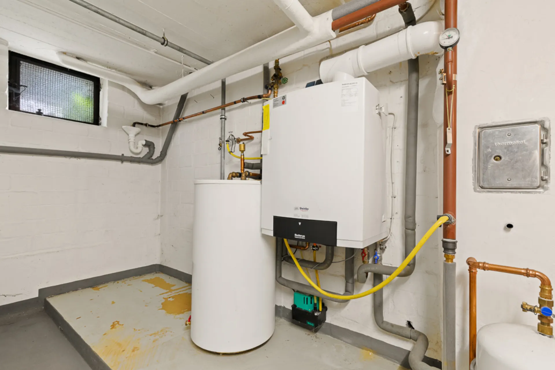 Heizungskeller mit Wandheizung, Rohren und Boiler, Fenster oben links an der Wand.