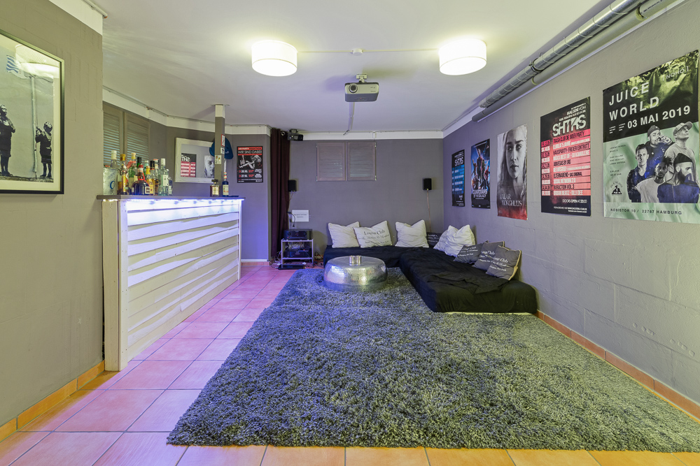 Wohnzimmer mit Bar, Teppich, niedrigen Sofas, vielen Kissen und Postern an den Wänden.