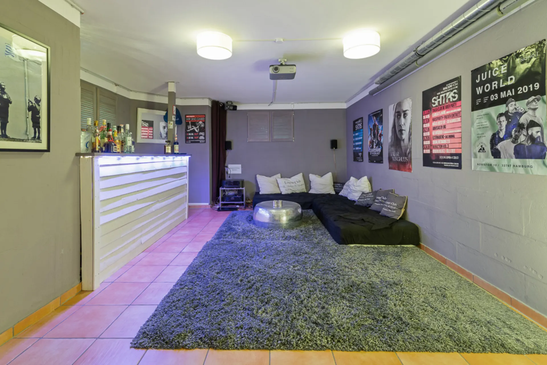 Wohnzimmer mit Bar, Teppich, niedrigen Sofas, vielen Kissen und Postern an den Wänden.