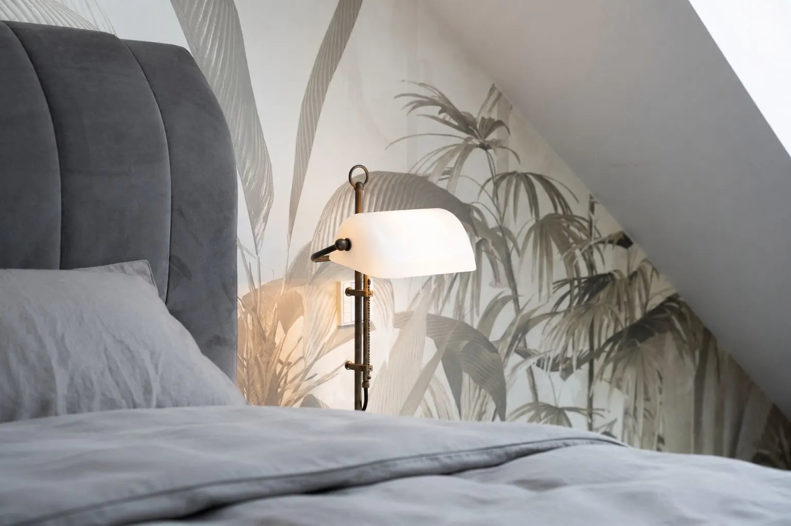 Teil eines Bettes mit grauer Bettwäsche, Nachttischlampe und Tapete mit Pflanzenmotiv.