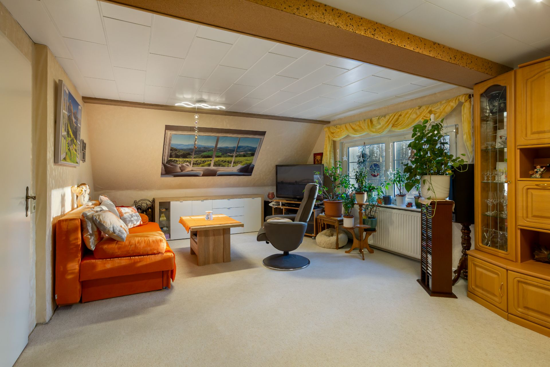Wohnzimmer mit orangefarbenem Sofa, Couchtisch, Sessel, großen Pflanzen und Fernseher am Fenster.