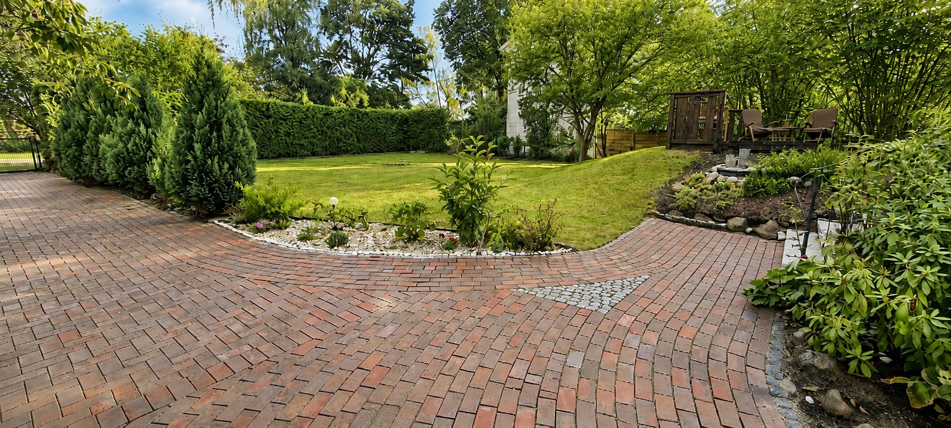 Gepflasterter Weg mit Garten, Rasenfläche, Büschen und Sitzbereich auf einer kleinen Anhöhe.