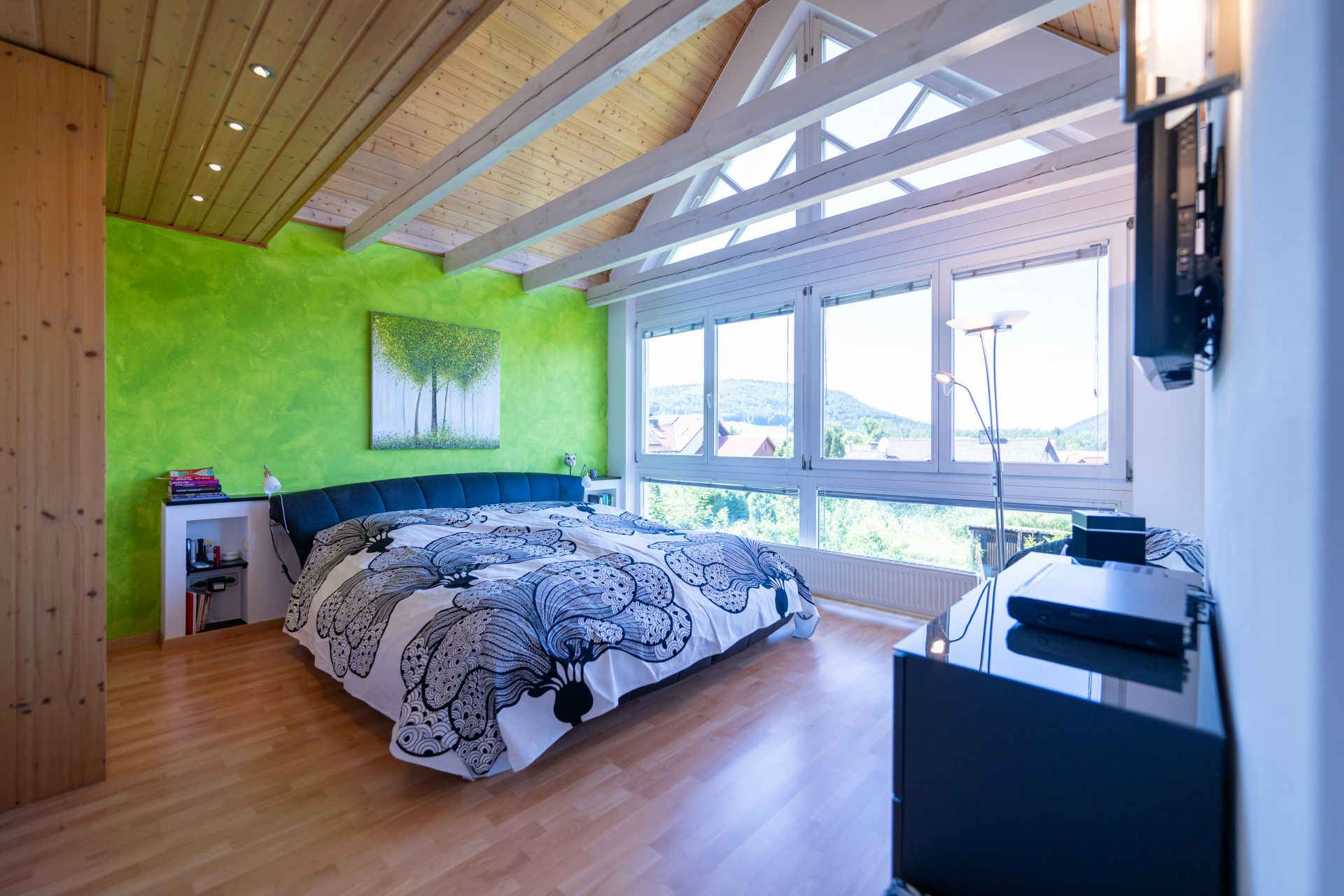 Helles Schlafzimmer mit großem Bett, grüner Wand und großen Fenstern mit Ausblick.