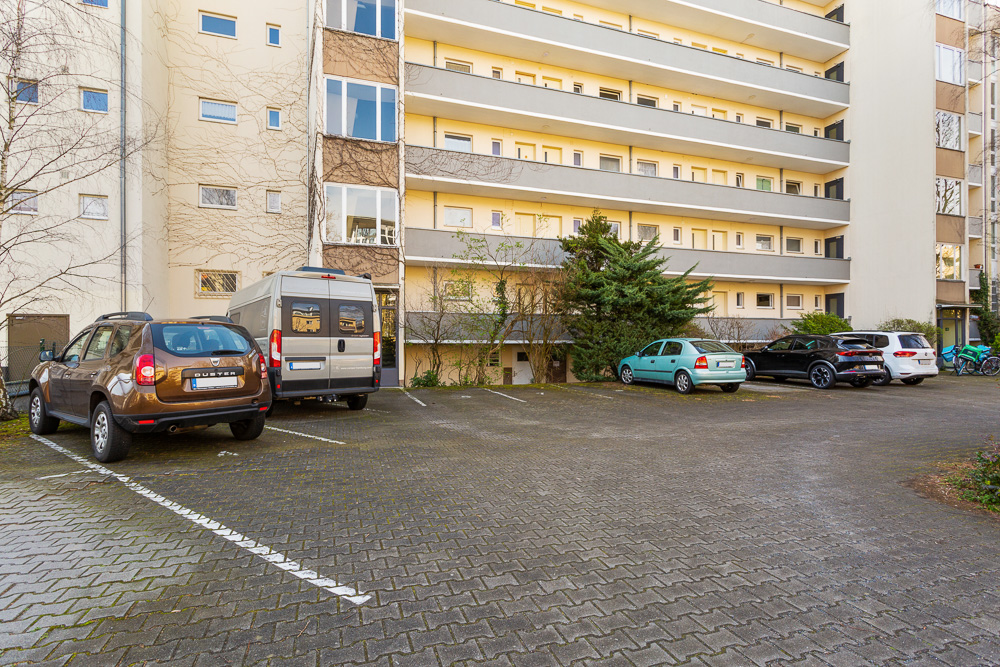 Mehrere Autos parken auf einem Parkplatz vor einem Wohngebäude mit Balkonen.