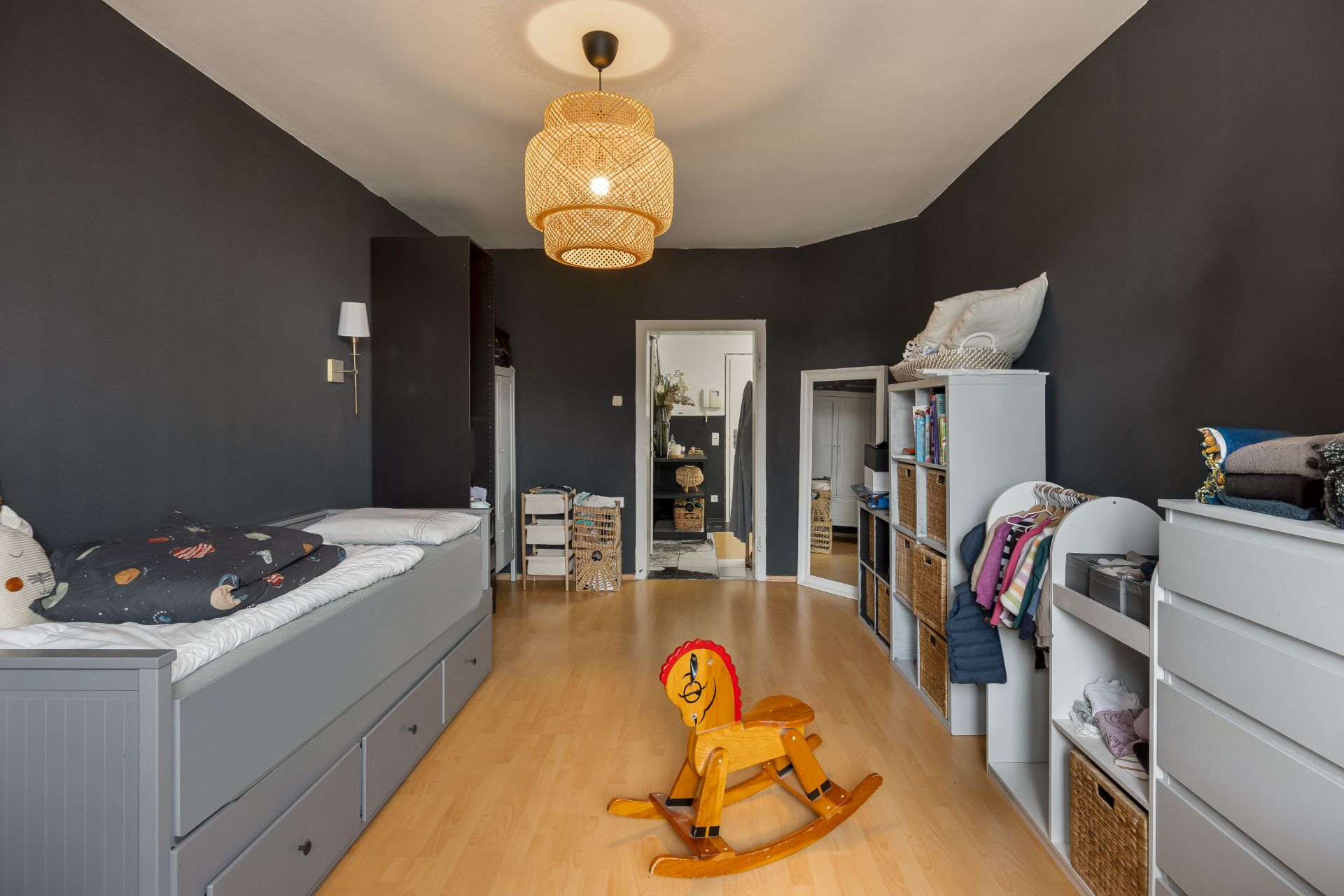 Kinderzimmer mit grauen Wänden, Bett, Regalen und Schaukelpferd aus Holz.
