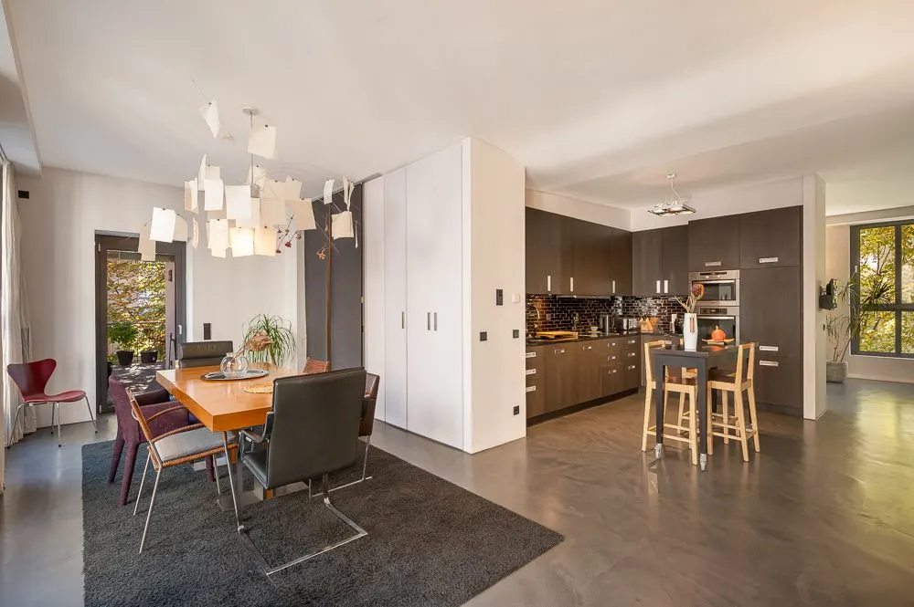 Moderne offene Küche mit Essbereich, Holztisch und Designerlampen, große Fenster im Hintergrund.